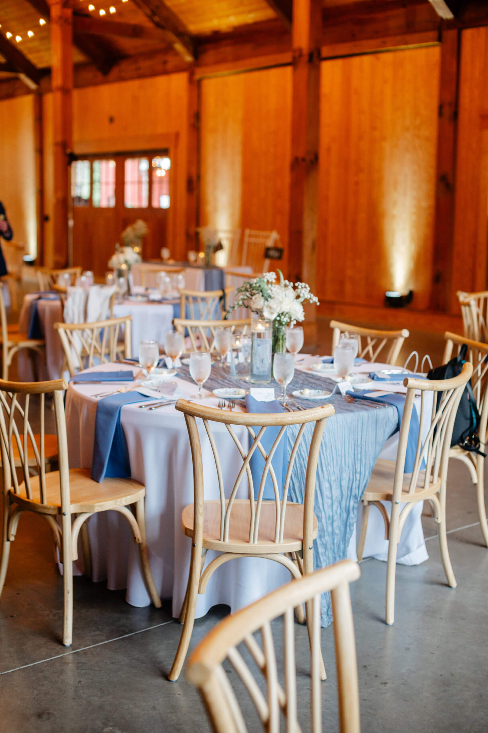 Barns of Kanak October Wedding 31 Round table set up in reception area with white table clothe, light blue runner and napkins, white dishes and a blue candle centerpiece with white flowers