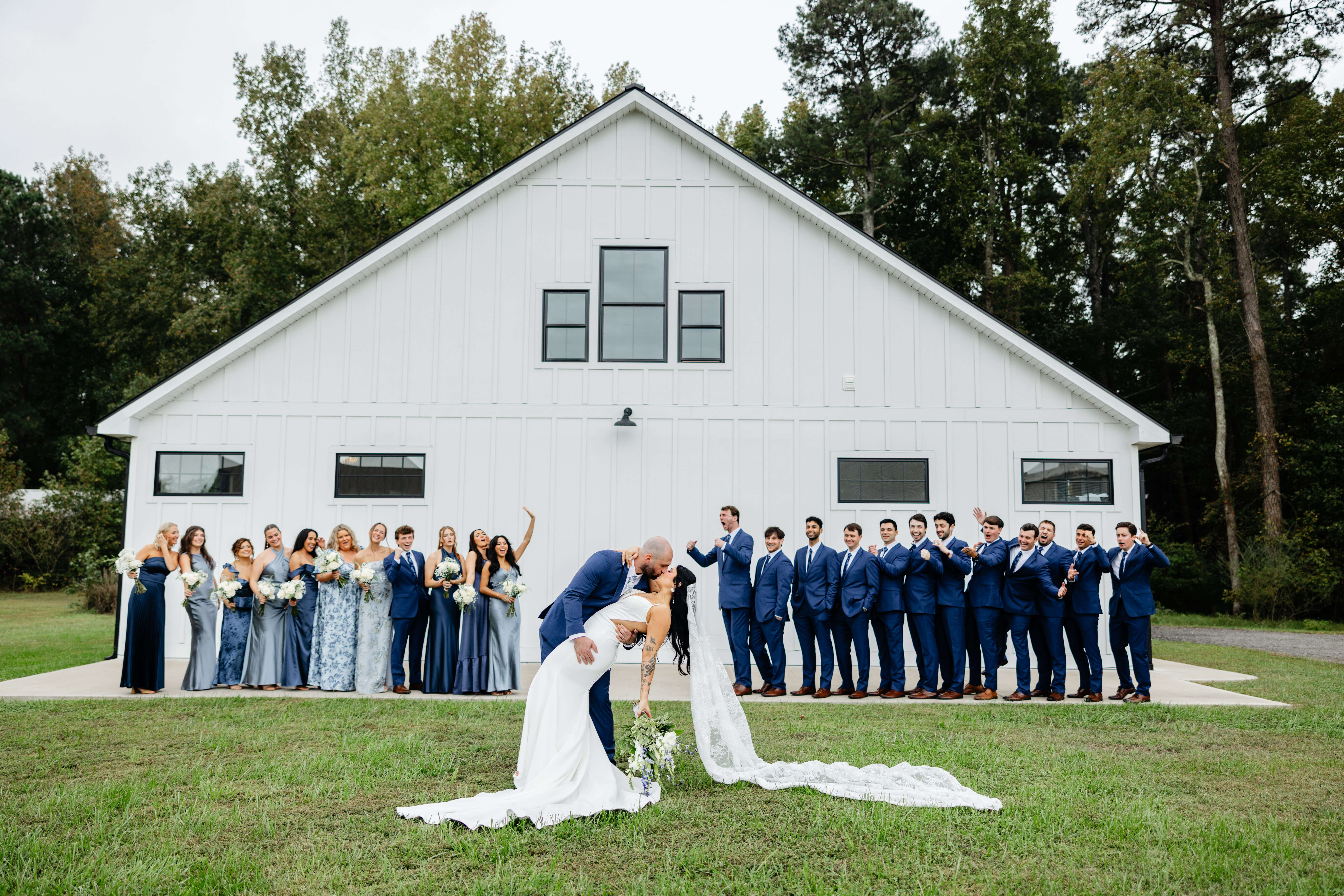 Barns of Kanak October Wedding 34 Groom leaning bride back for a kiss outside as wedding party cheer in background, everyone in front of white barn