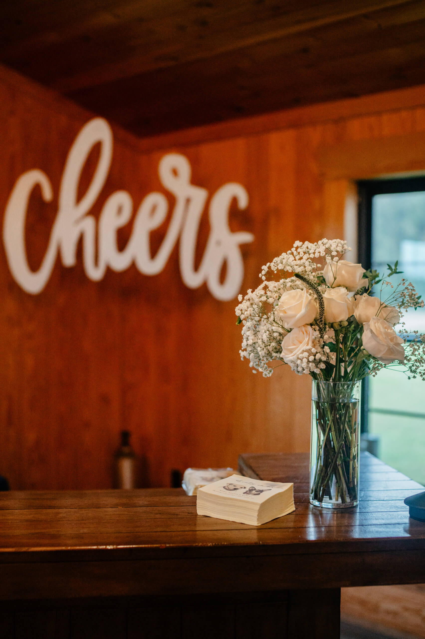 Barns of Kanak October Wedding 35 Glass vase of white flowers sits on corner of bar in reception space, 'Cheers' written on wall in background, and stack of napkins on bar with bride and groom's pets on them