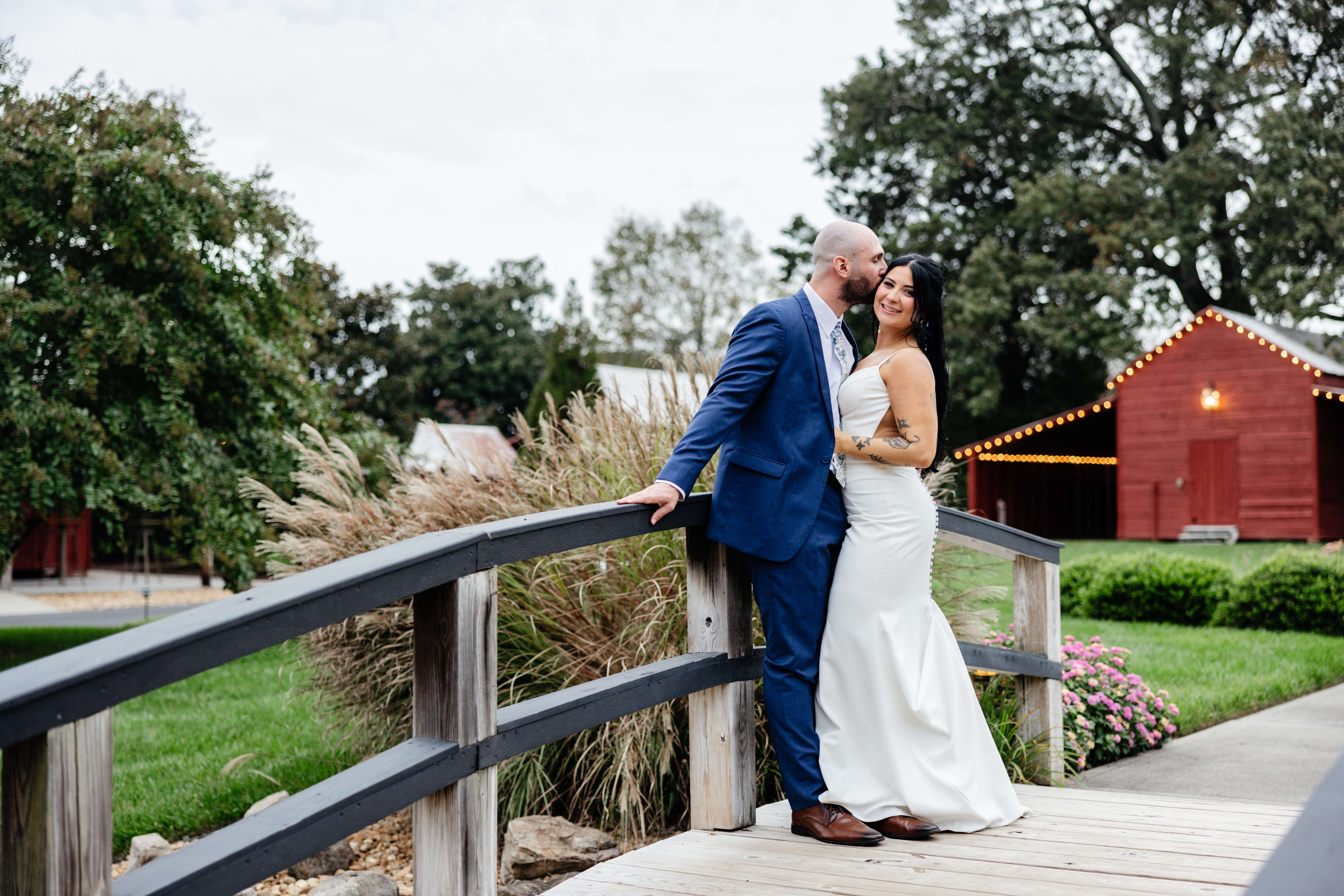Barns of Kanak October Wedding 44 Groom kisses bride's head as she smiles at camera, both standing on small bridge outside of venue, red barn in background