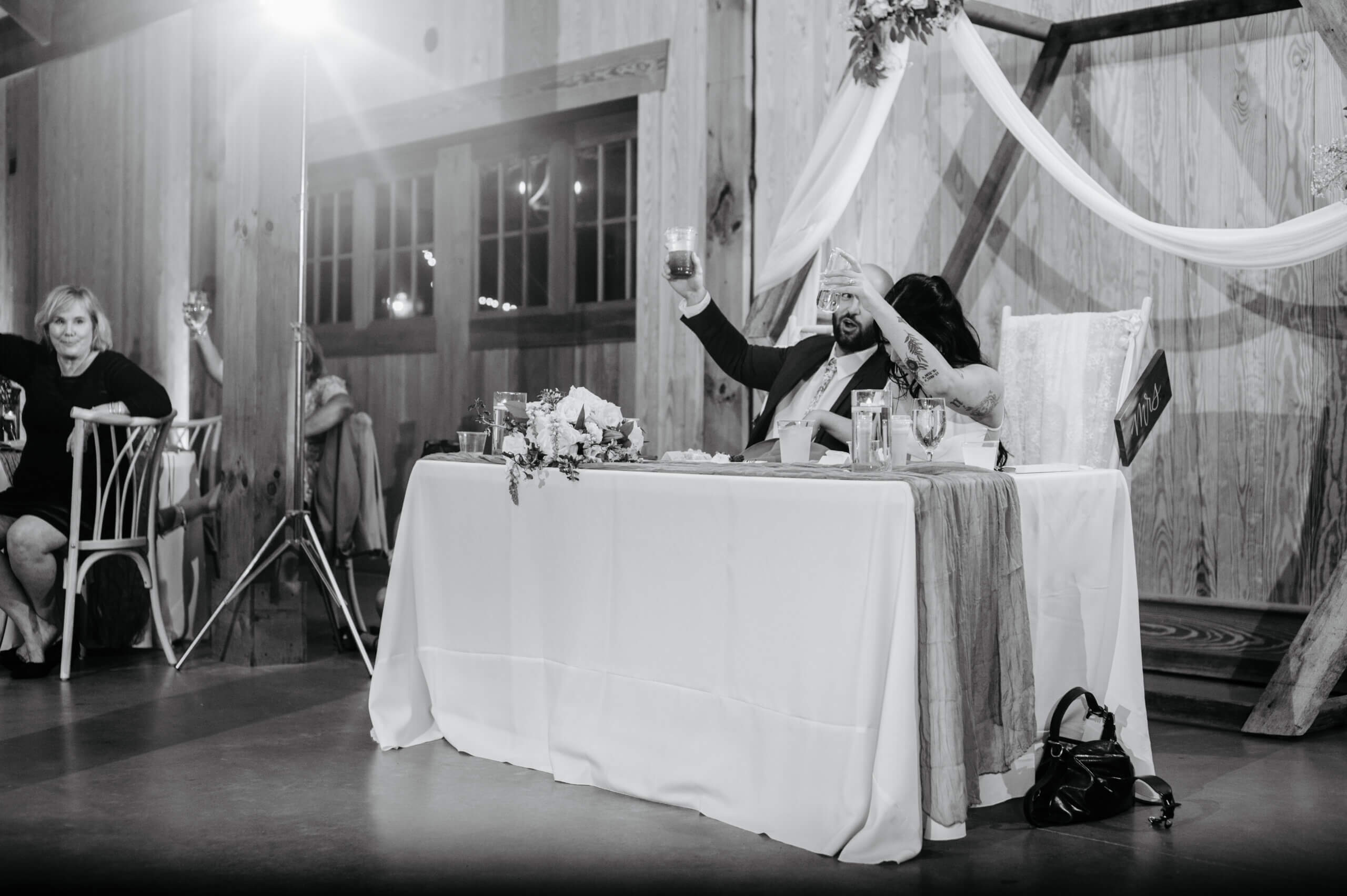 Barns of Kanak October Wedding 51 Black and white photo of bride and groom holding up their glasses as they smile while sitting at their sweetheart table