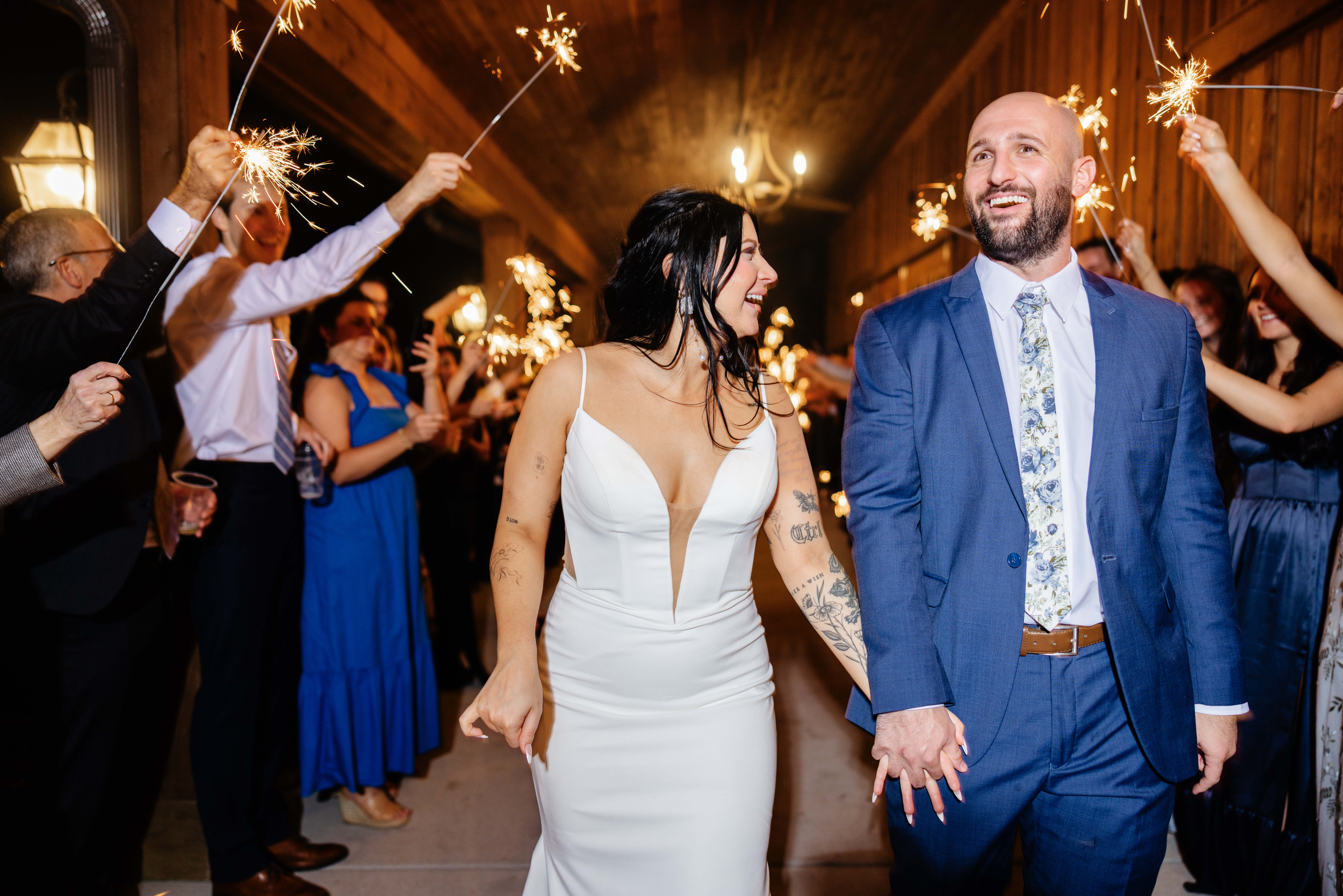 Barns of Kanak October Wedding 57 Bride and groom holding hands, bride smiling at groom as he looks forwards, guests holding sparklers on either side and in background