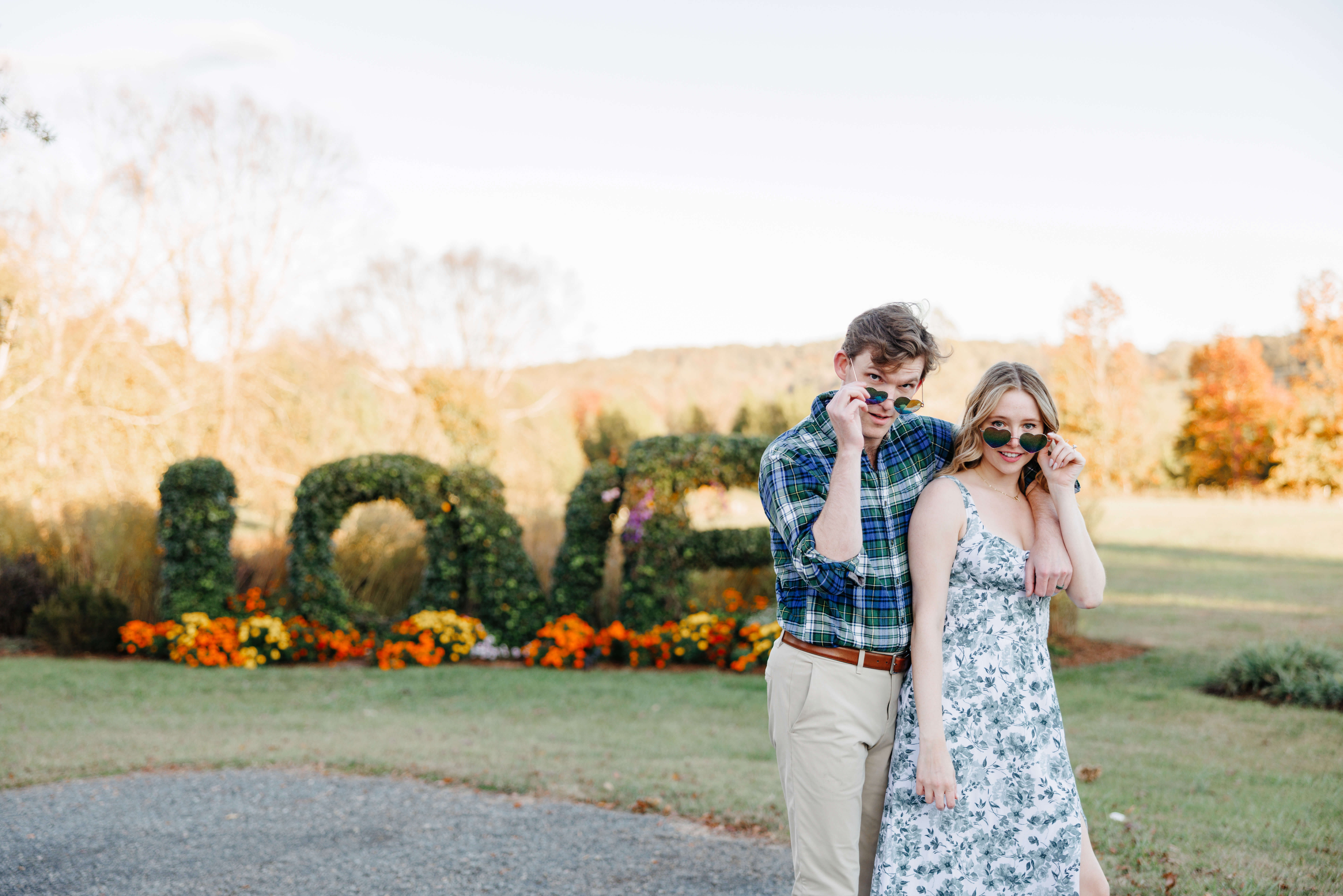 Market at Grelen Fall Engagement Photography 18 Engaged couple striking a pose with heart shaped sunglasses they're pulling down their noses to look at the camera over them, LOVE in plants spelled out behind them
