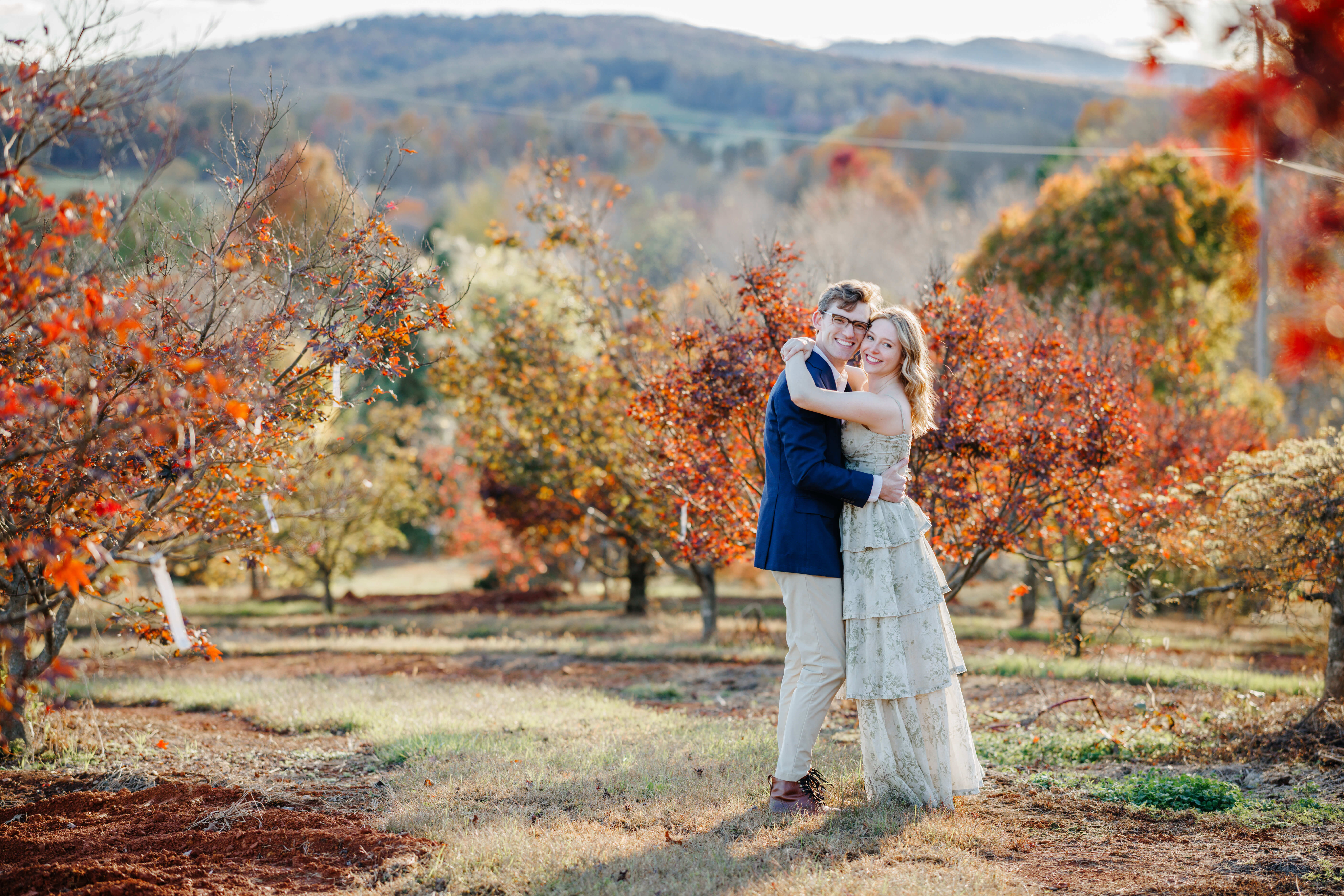 Market at Grelen Fall Engagement Photography 2 Man and woman with their arms around one another stand amongst the fall colored trees as they both smile at the camera