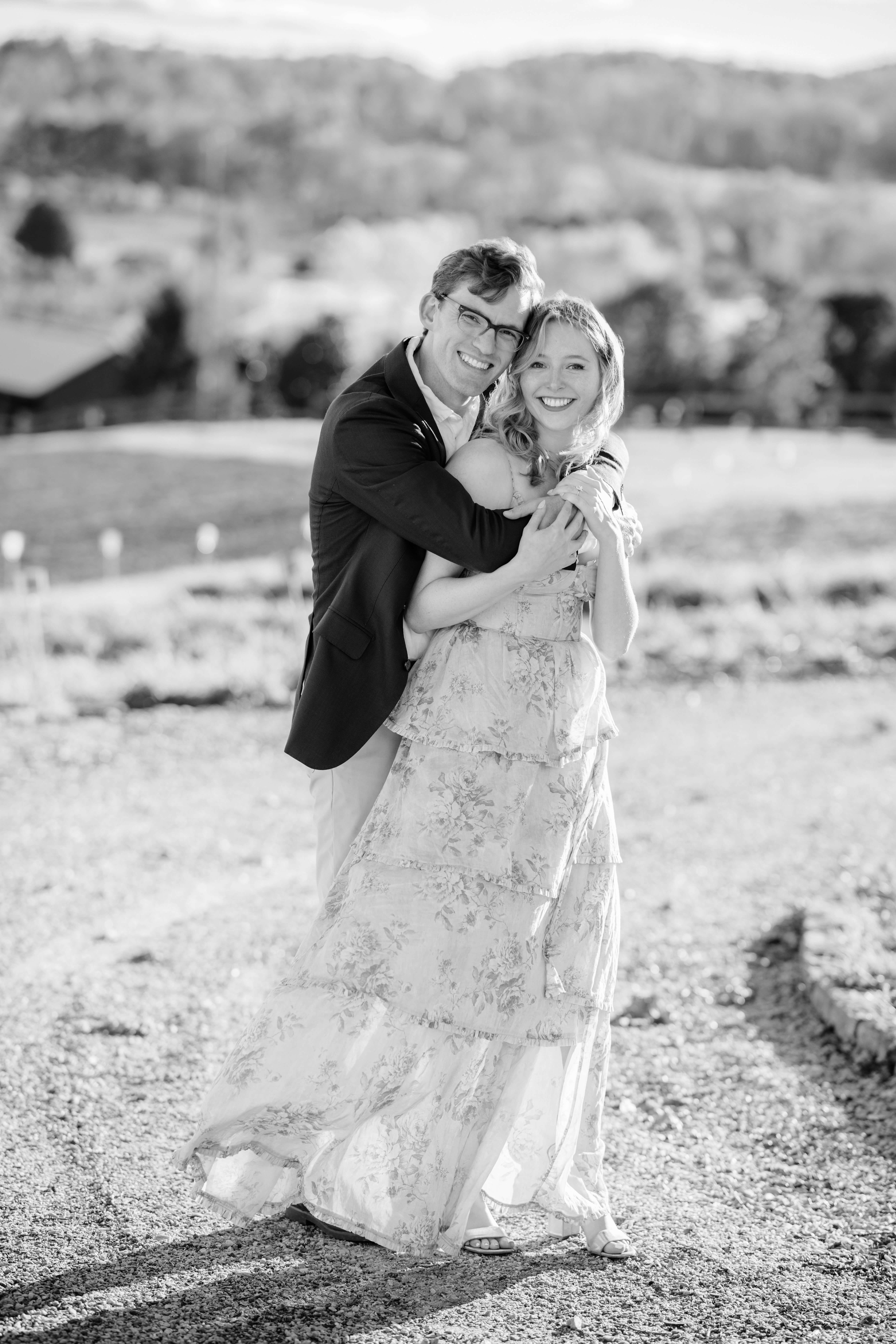 Market at Grelen Fall Engagement Photography 6 Black and white photo of engaged couple standing outside on a dirt road at market at grelen, man hugging woman from behind as they both smile at the camera