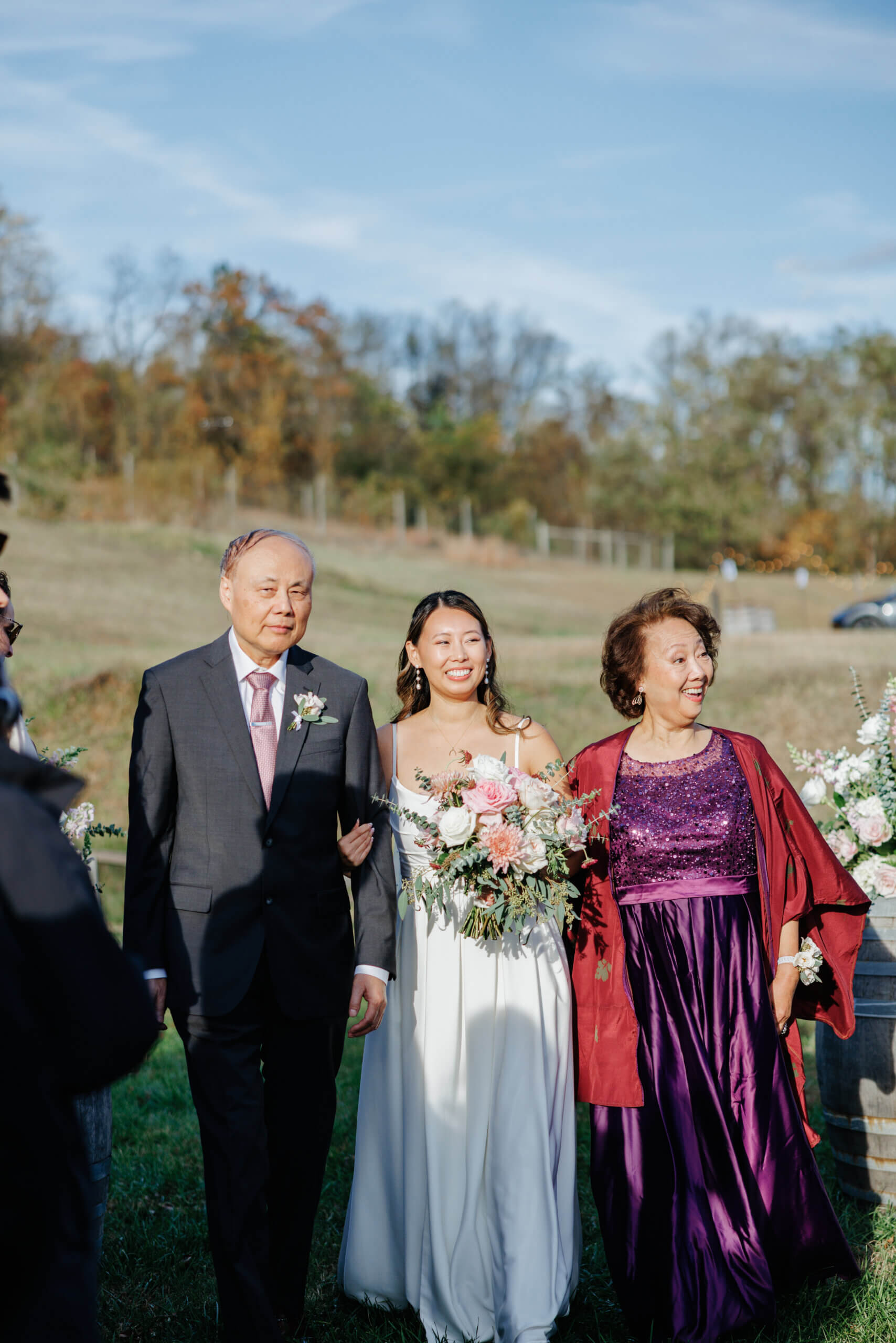 Valley Road Vineyard Fall Wedding 22 Bride walks down aisle arm in arm with her parents, her mom with an excited smile on her face