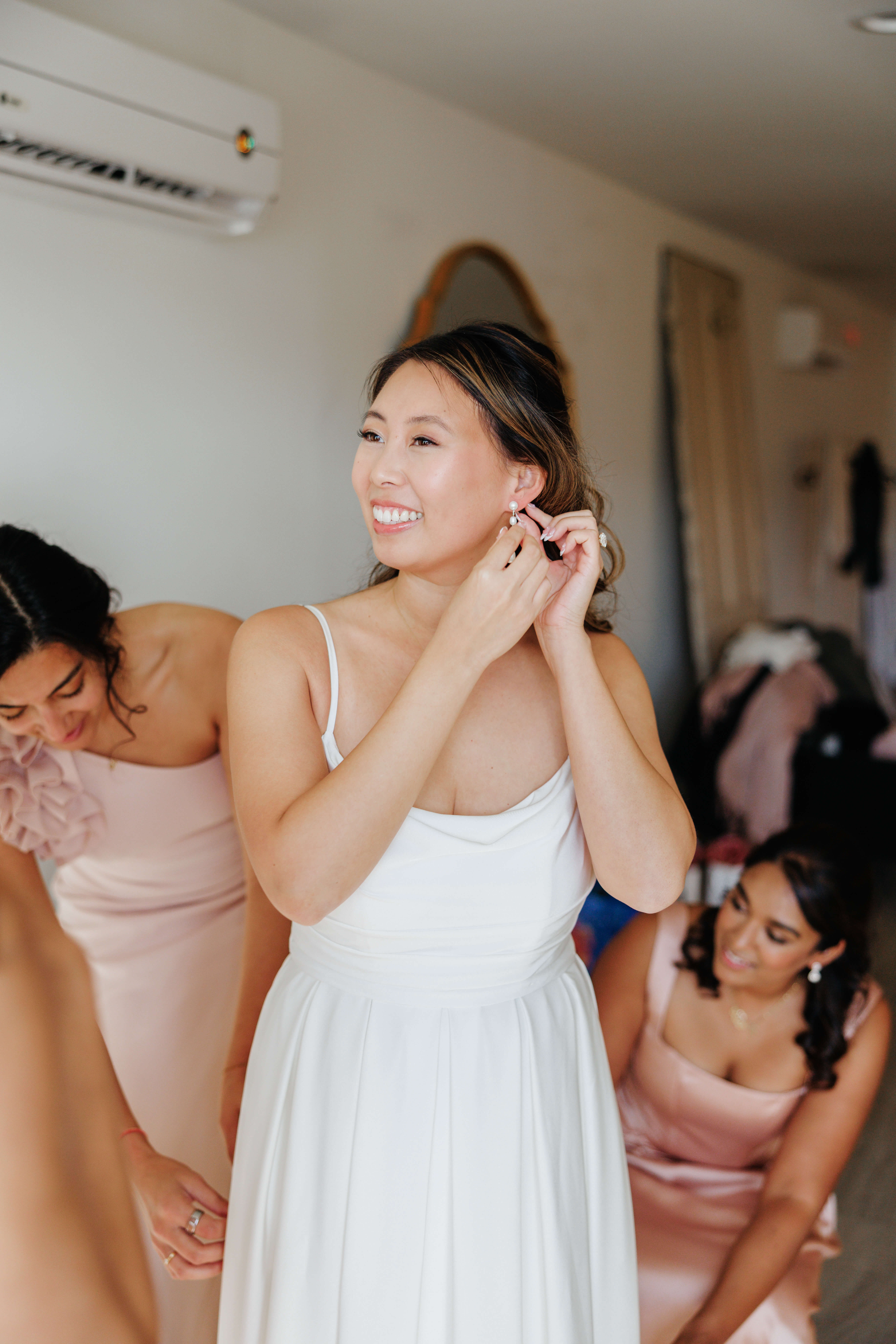 Valley Road Vineyard Fall Wedding 5 Bride in her wedding dress putting in a white earring as her bridesmaids in light pink dresses adjust her dress around her