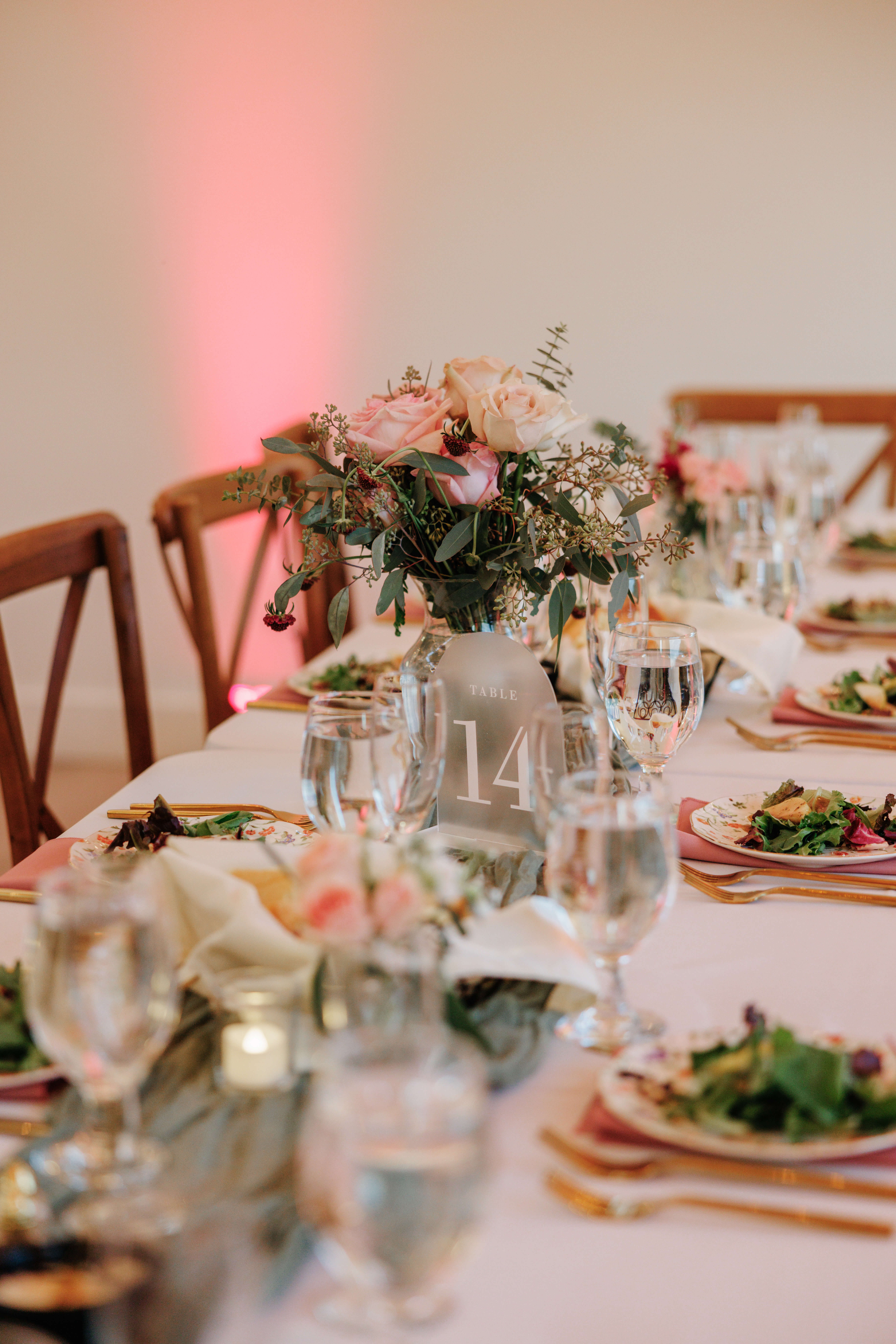 Valley Road Vineyard Fall Wedding 37 Table centerpiece of pink flowers and frosted glass table number sign sit on white table in the reception space, pink up lighting in background
