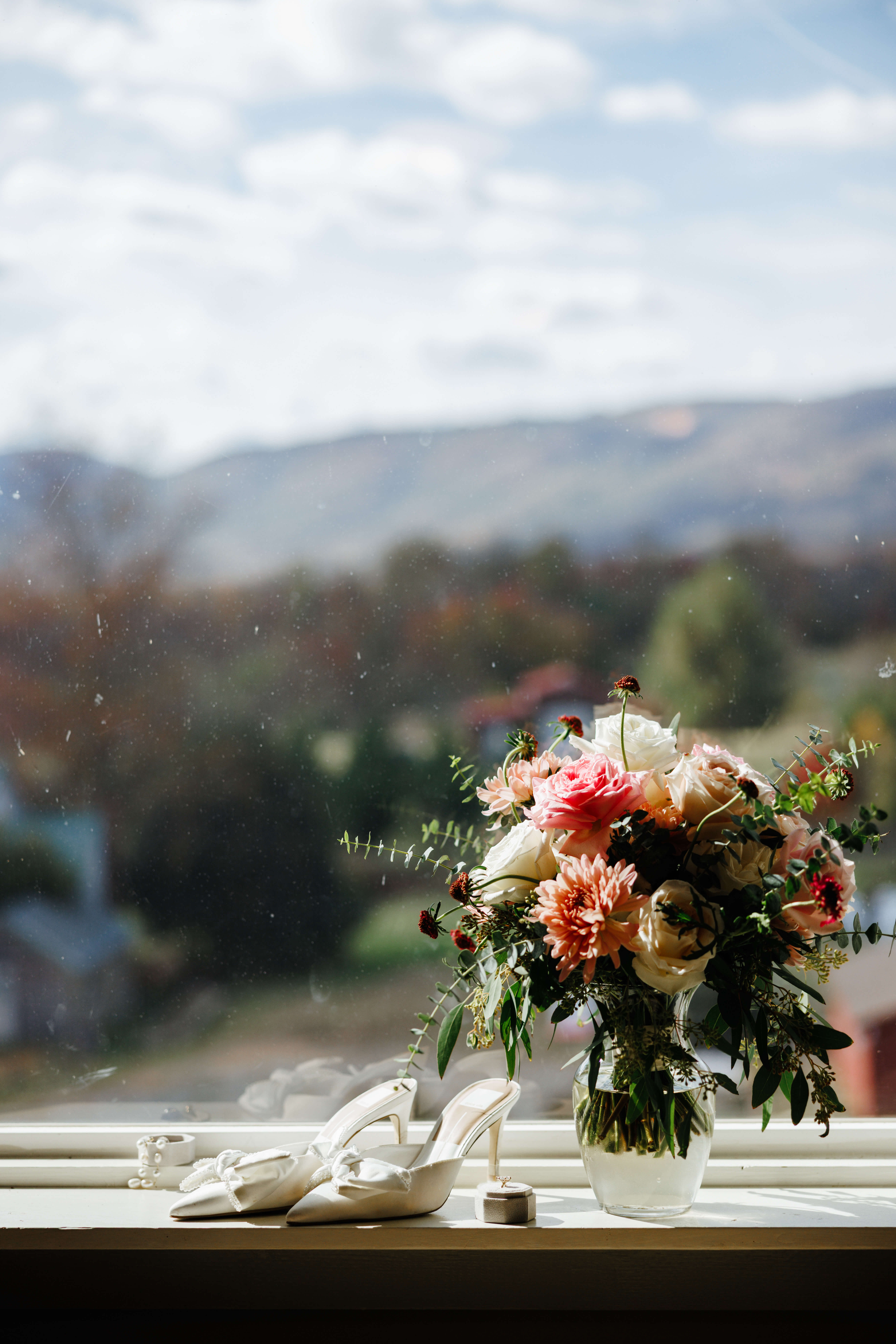 Valley Road Vineyard Fall Wedding 1 Bride's bouquet of white and pink flowers sits in a clear vase on windowsill next to her white wedding shoes, outside window is beautiful mountain view