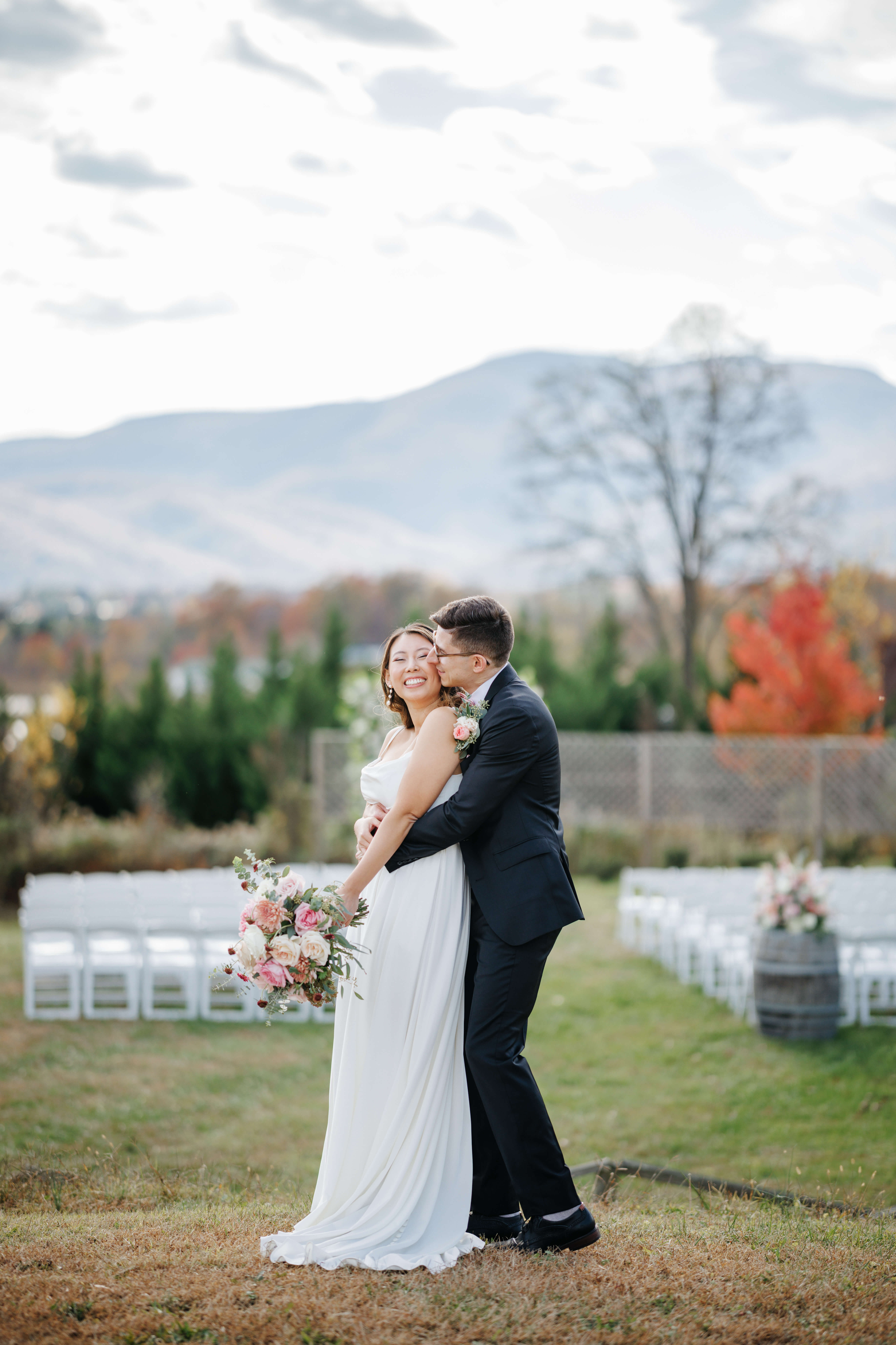Valley Road Vineyard Fall Wedding 9 Groom hugging bride from behind and kissing her cheek as she smiles at camera and holds her bouquet of pink flowers