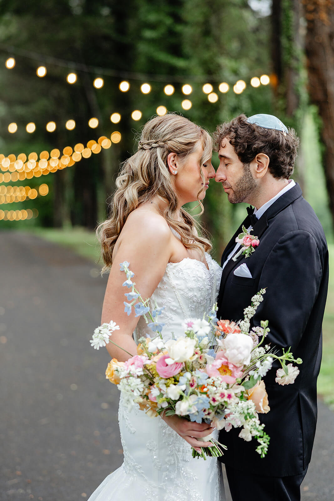 Poplar Springs Manor Spring Wedding Photography 26 porttrait of bride and groom touching noses, they are standing by a wooded path with string lights in the background