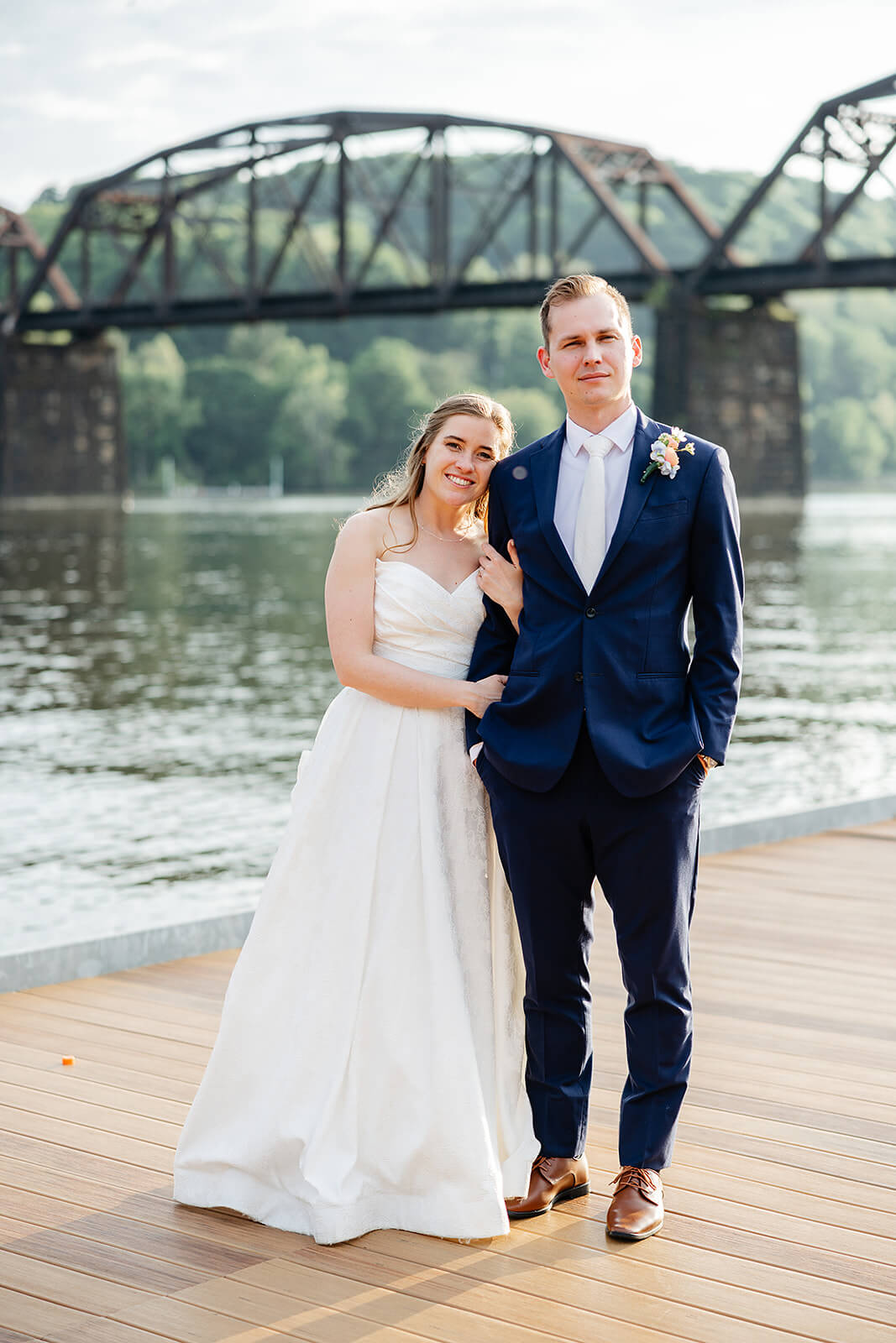 Allegheny RiverTrail Park Spring Wedding Photography 39 bride hugging grooms arm as they stand side by side, they are on a riverside dock with a bridge in the background
