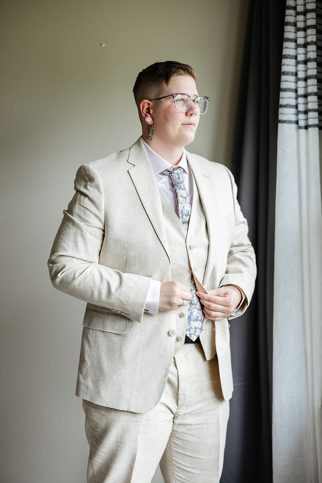 Vaughan Greenhouse Spring Wedding Photography 4 groom buttoning jacket of tan suit in light of a window