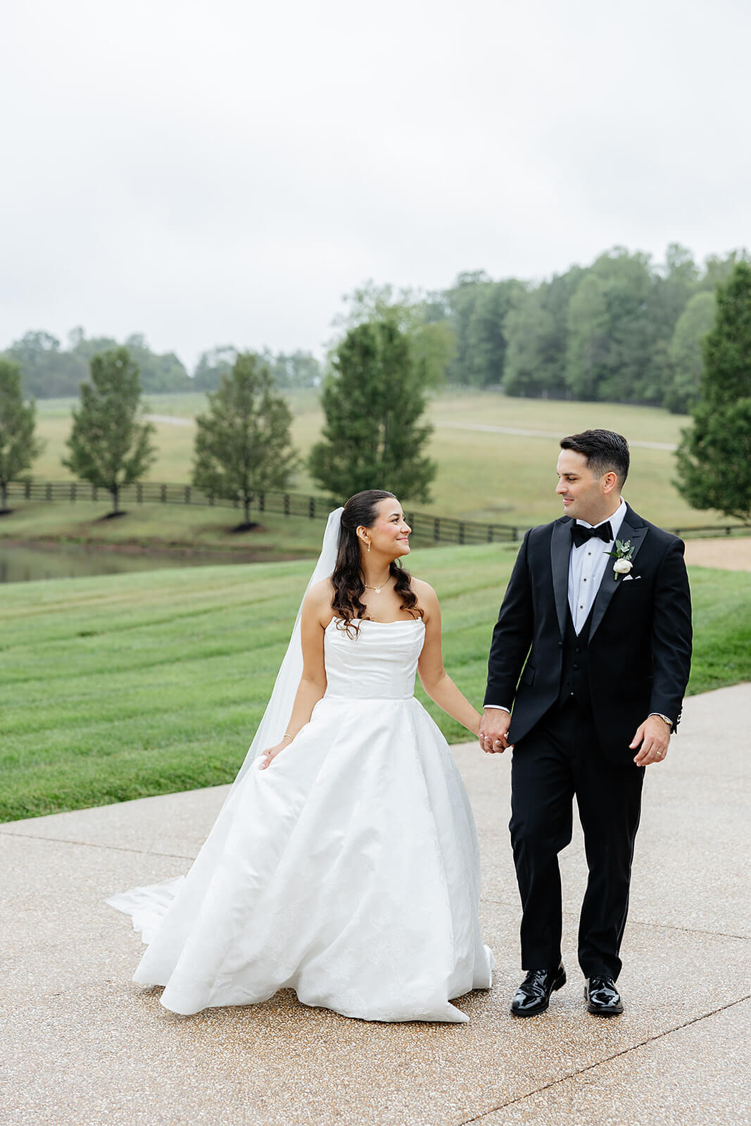 Matt + Adriana's May Wedding Photography 21 bride and groom holding hands outside on a path, there is a field behind them that is wet from rain