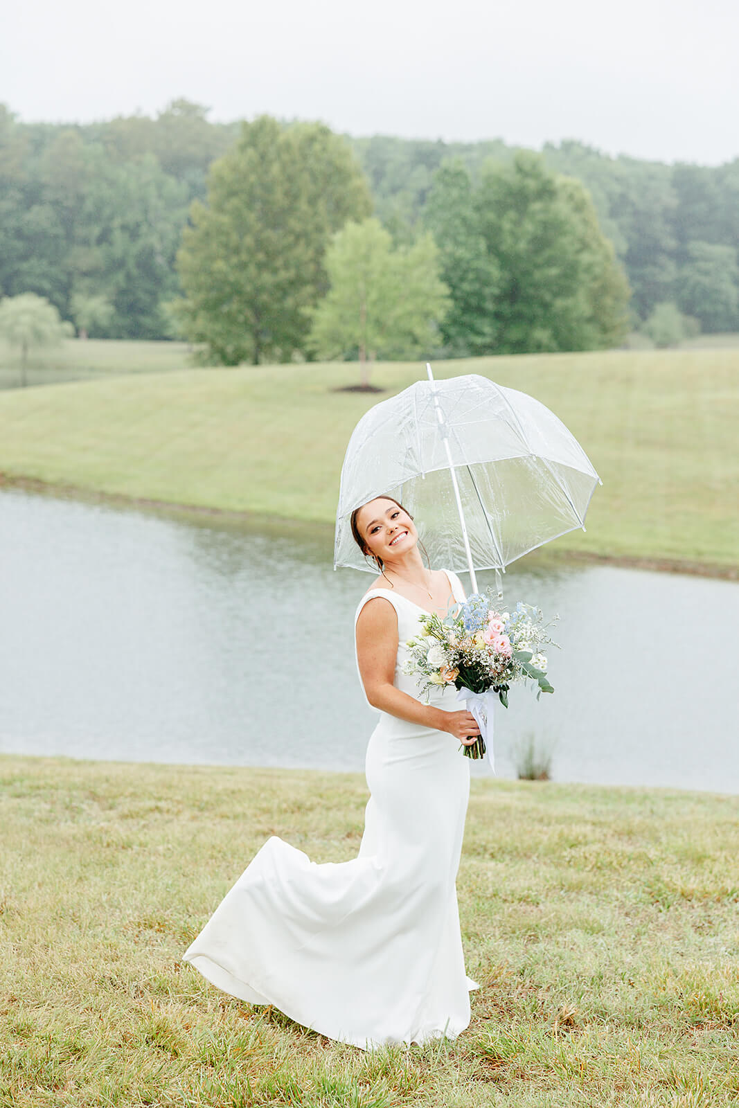 Family Farm Spring Wedding Photography 23 bride in front of pond kicking her foot up and holding a clear umbrella