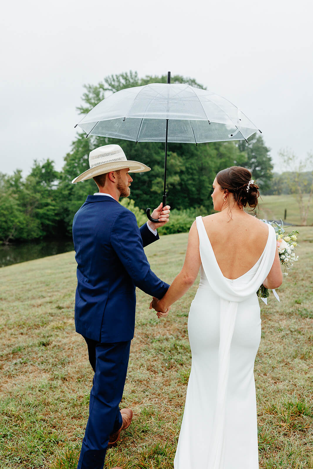 Family Farm Spring Wedding Photography 26 bride and groom holding hands and walking up a hill, groom is holding an umbrella over them