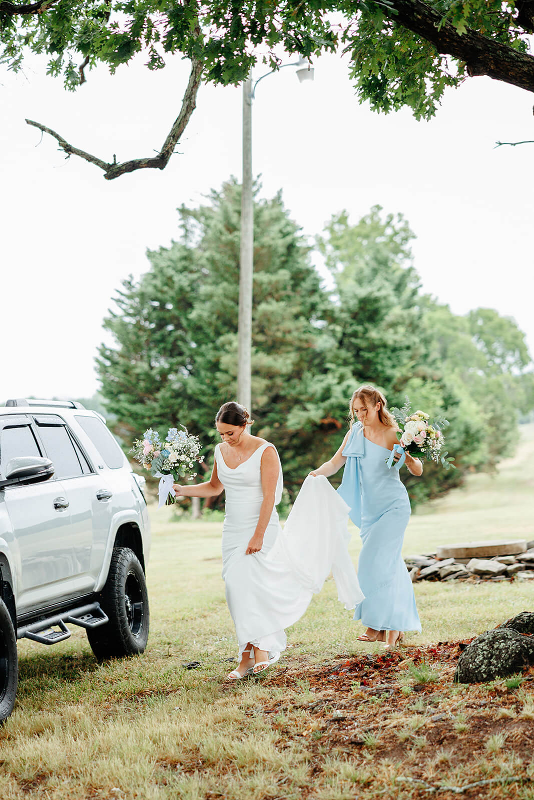 Family Farm Spring Wedding Photography 8 bridesmaid helping bride carry her dress and walk across a field