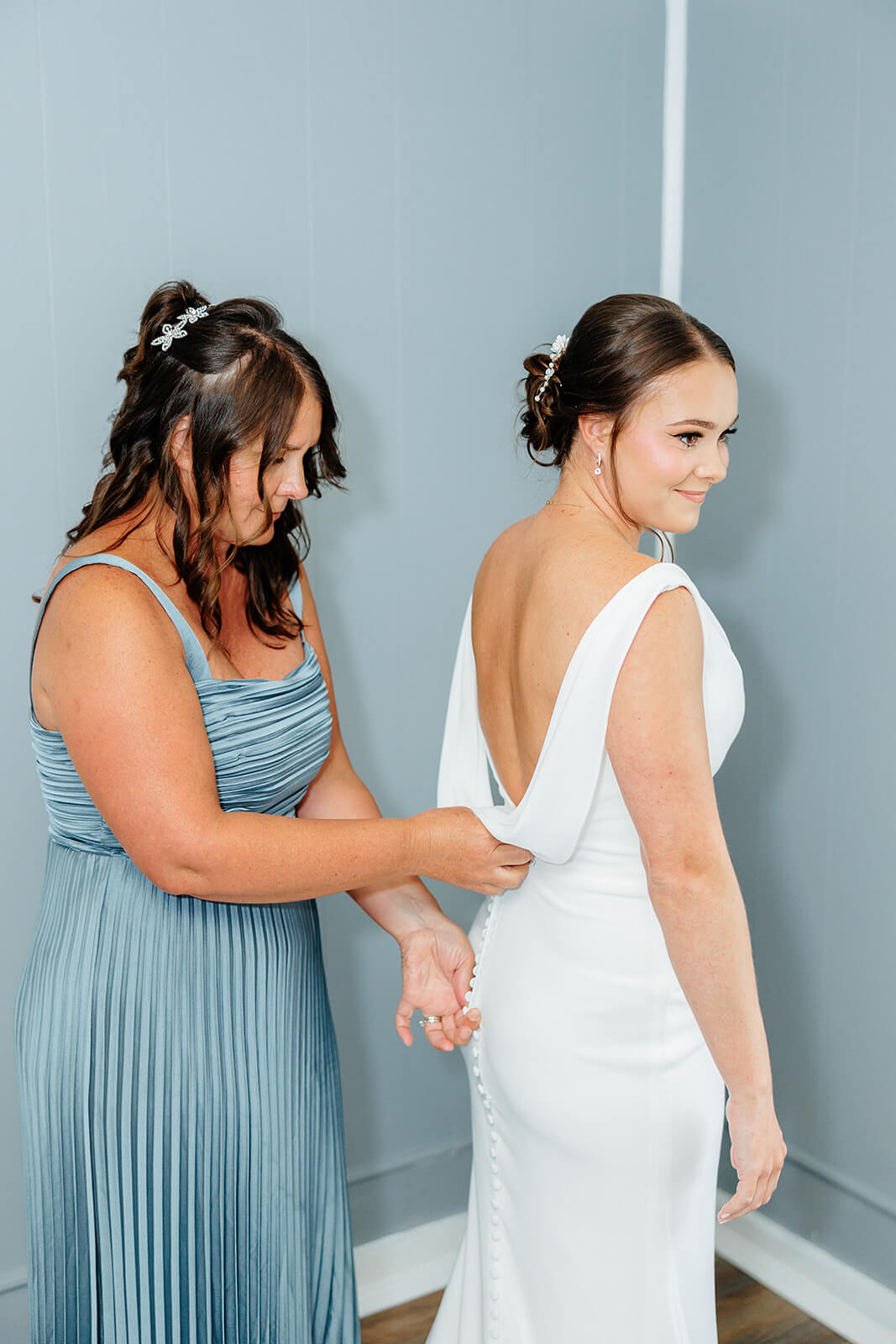Family Farm Spring Wedding Photography 2 bridesmaid in blue dress helping bride button her wedding dress