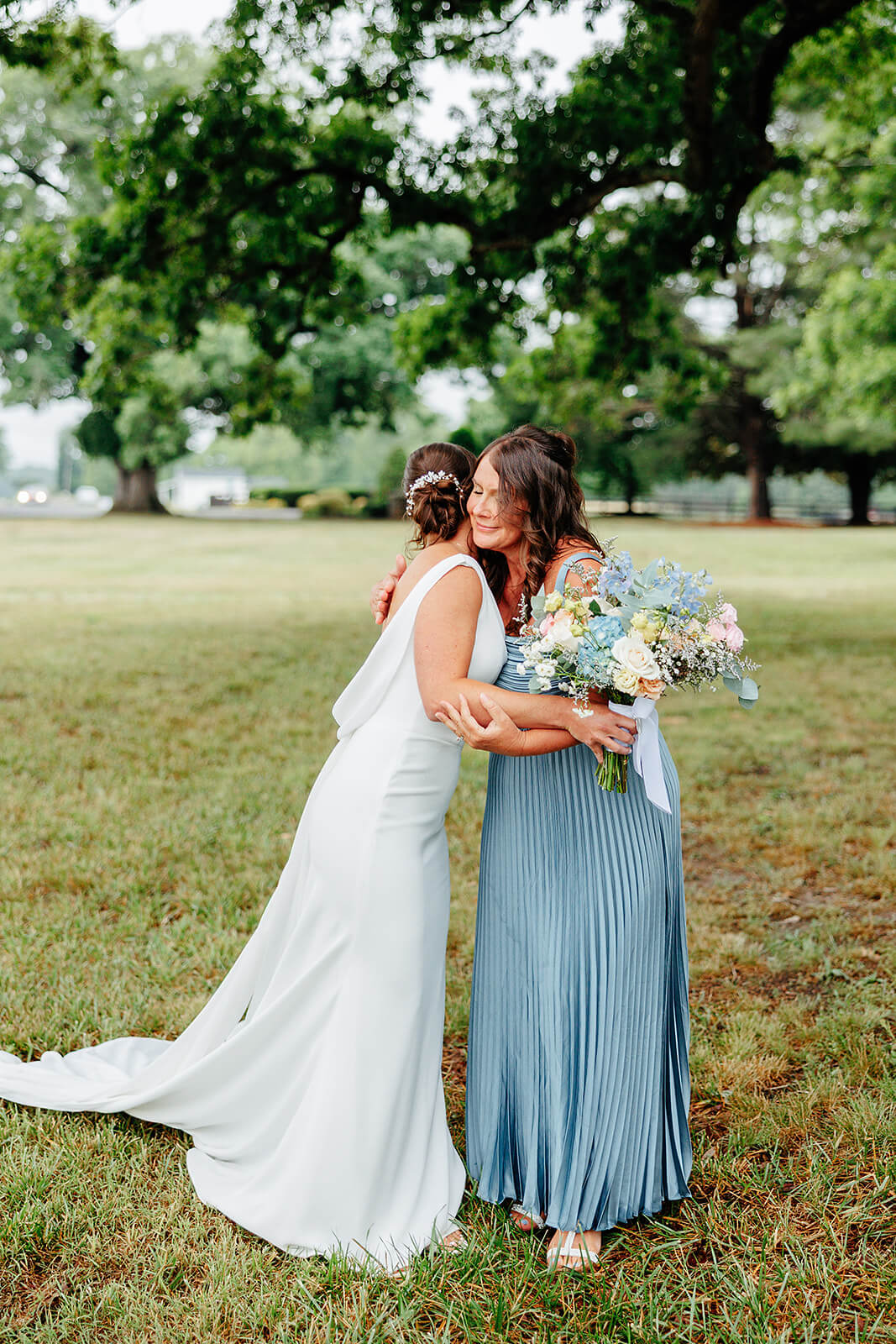 Family Farm Spring Wedding Photography 11 bride hugging a bridesmaid in a field with trees in the near background
