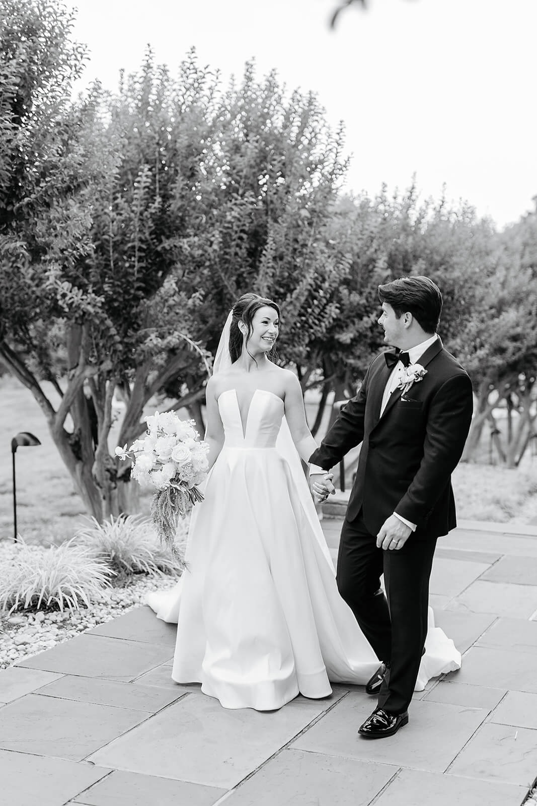Estate at River Run Summer Wedding Photography 22 black and white photo of bride and groom holding hands and walking down a stone path between crepe myrtle trees