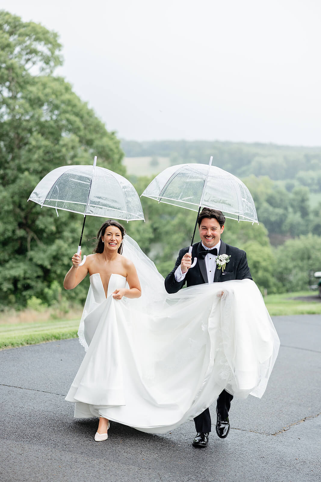 Estate at River Run Summer Wedding Photography 25 bride and groom walking in the rain with clear umbrellas, and the groom is holding the train of the brides dress