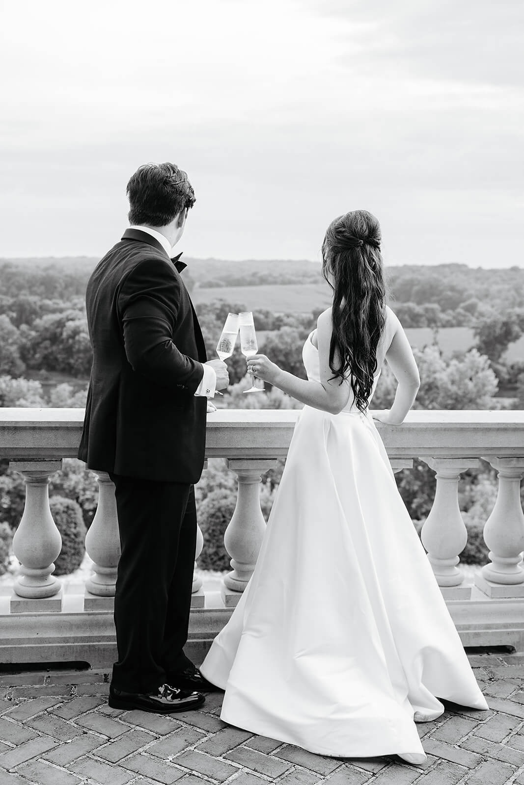 Estate at River Run Summer Wedding Photography 32 black and white photo of bride and groom facing away from the camera staring over a balcony and clinking champagne glasses