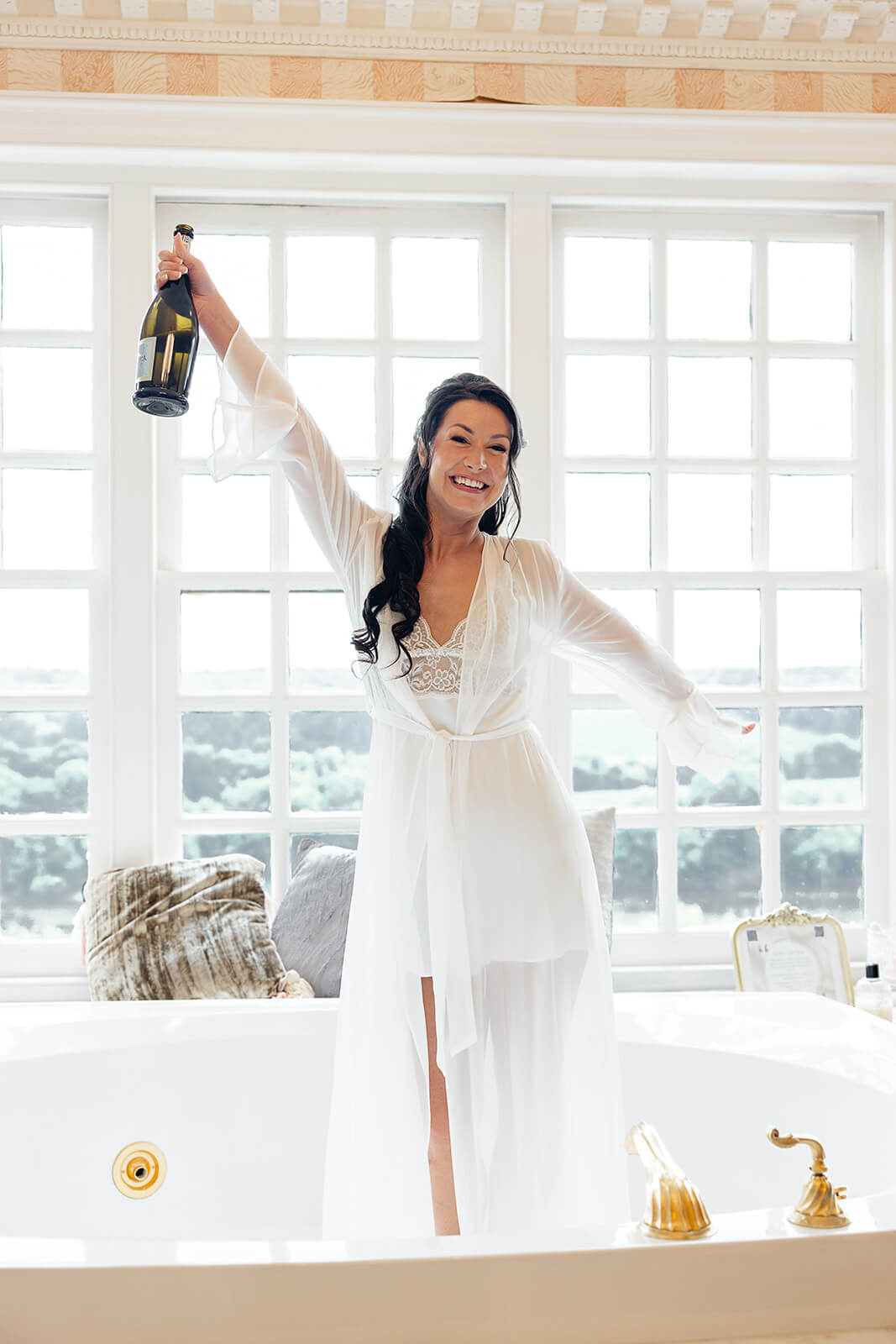 Estate at River Run Summer Wedding Photography 7 bride wearing white dressing robe and standing in the tub holding a bottle of champagne, there is a huge window behind her with lots of natural light