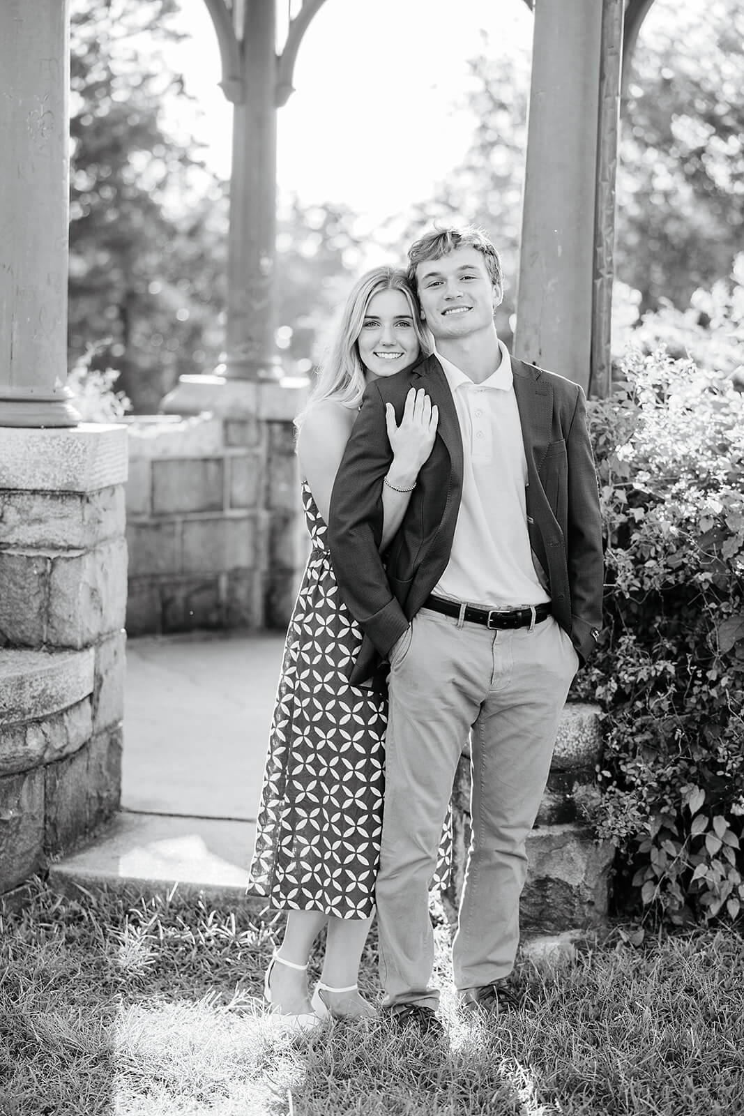 Maymont Summer Engagement Photography 15 black and white photo of woman hugging her fiance from behind as they stand in front of a gazebo with stone accents