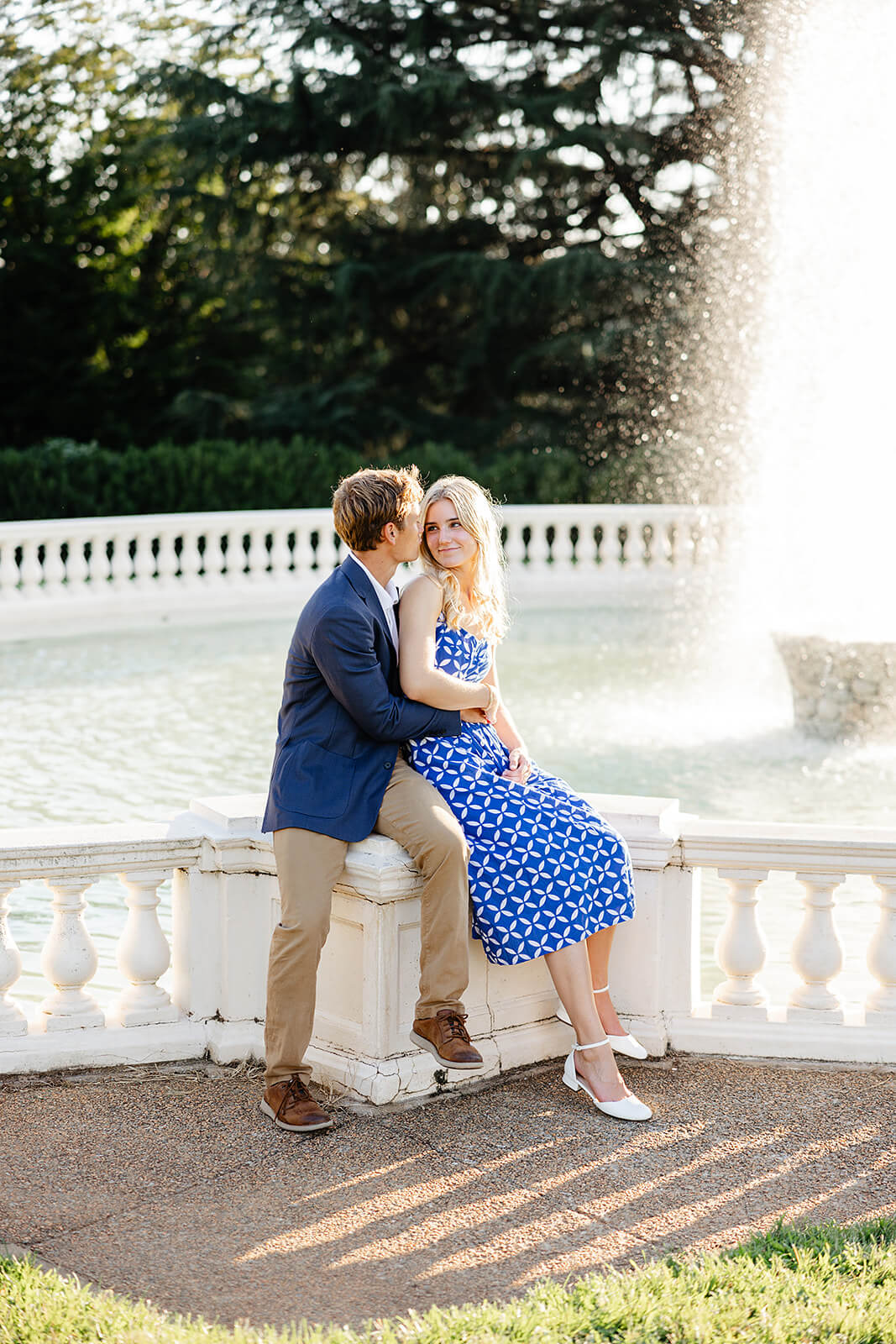 Maymont Summer Engagement Photography 16 couple sitting on edge of a fountain that is spurting water, he is kissing her cheek