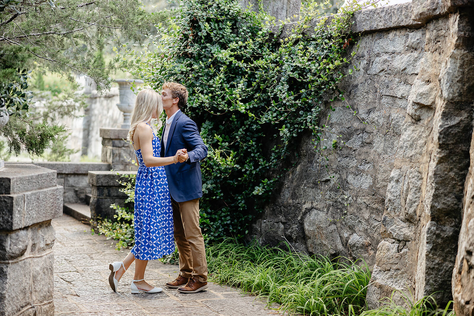 Maymont Summer Engagement Photography 3 man kisses woman's head in front of large stone wall with shrubbery around it