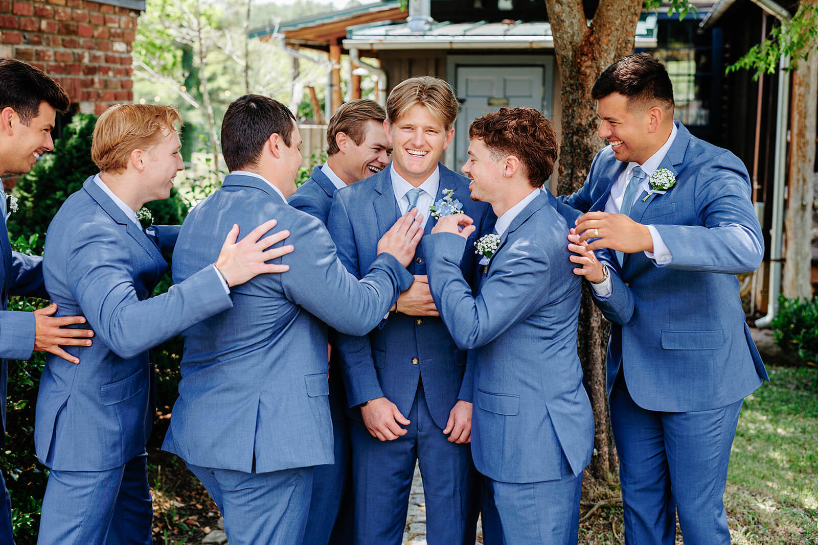 Rassawek Vineyard Spring Wedding Photography 2 groom with all his groomsmen standing around him and hugging him, they are wearing blue suits and outside by a large tree
