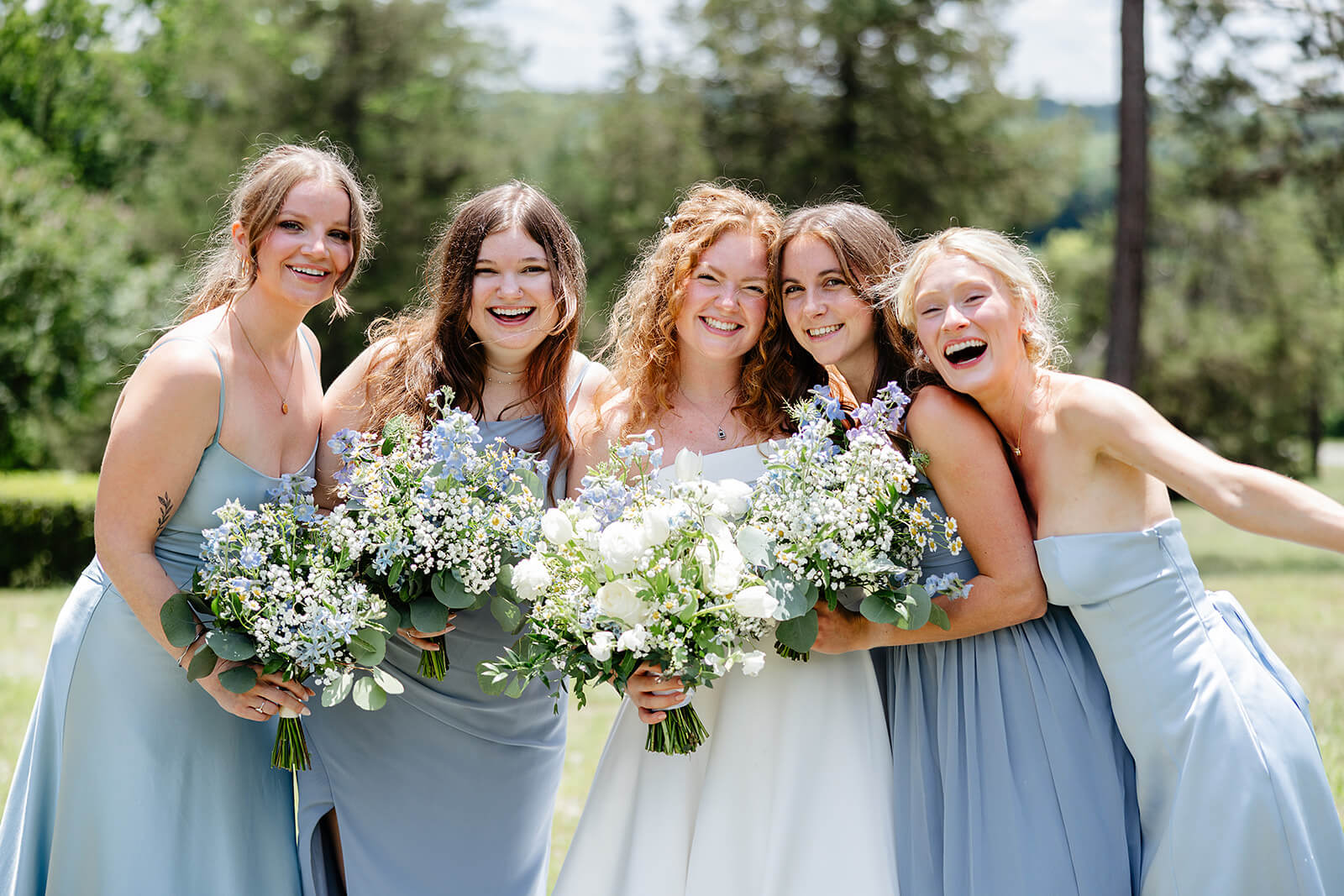 Rassawek Vineyard Spring Wedding Photography 9 bride and bridesmaid in blue dresses holding blue and white flowers and standing outside with trees in the background