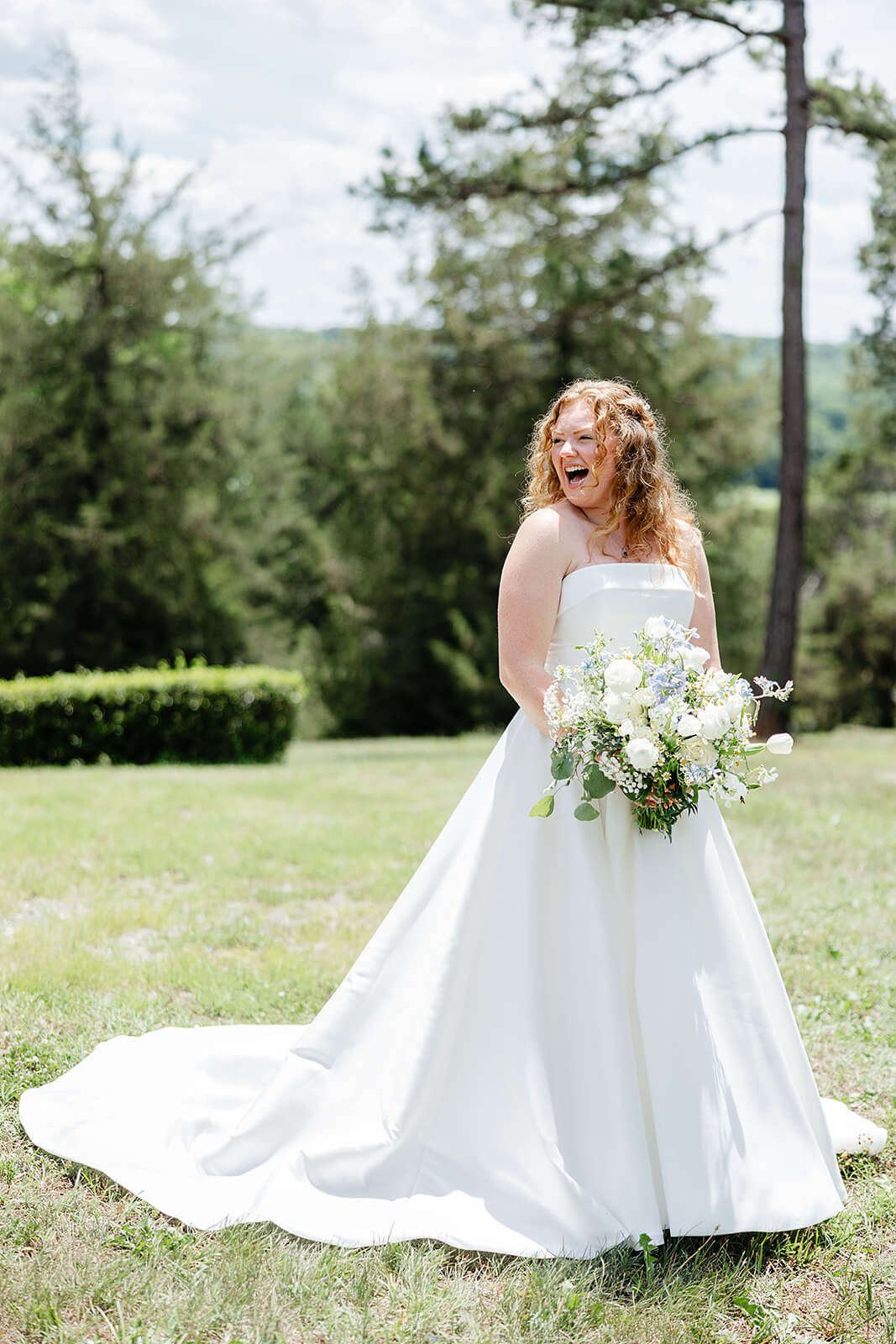 Rassawek Vineyard Spring Wedding Photography 10 bride in her dress and holding her bouquet standing on a lawn with trees and shrubs in the background