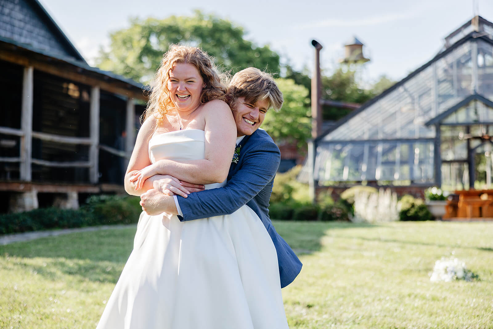 Rassawek Vineyard Spring Wedding Photography 27 groom hugging bride from behind, there is a large greenhouse and a large cabin the background