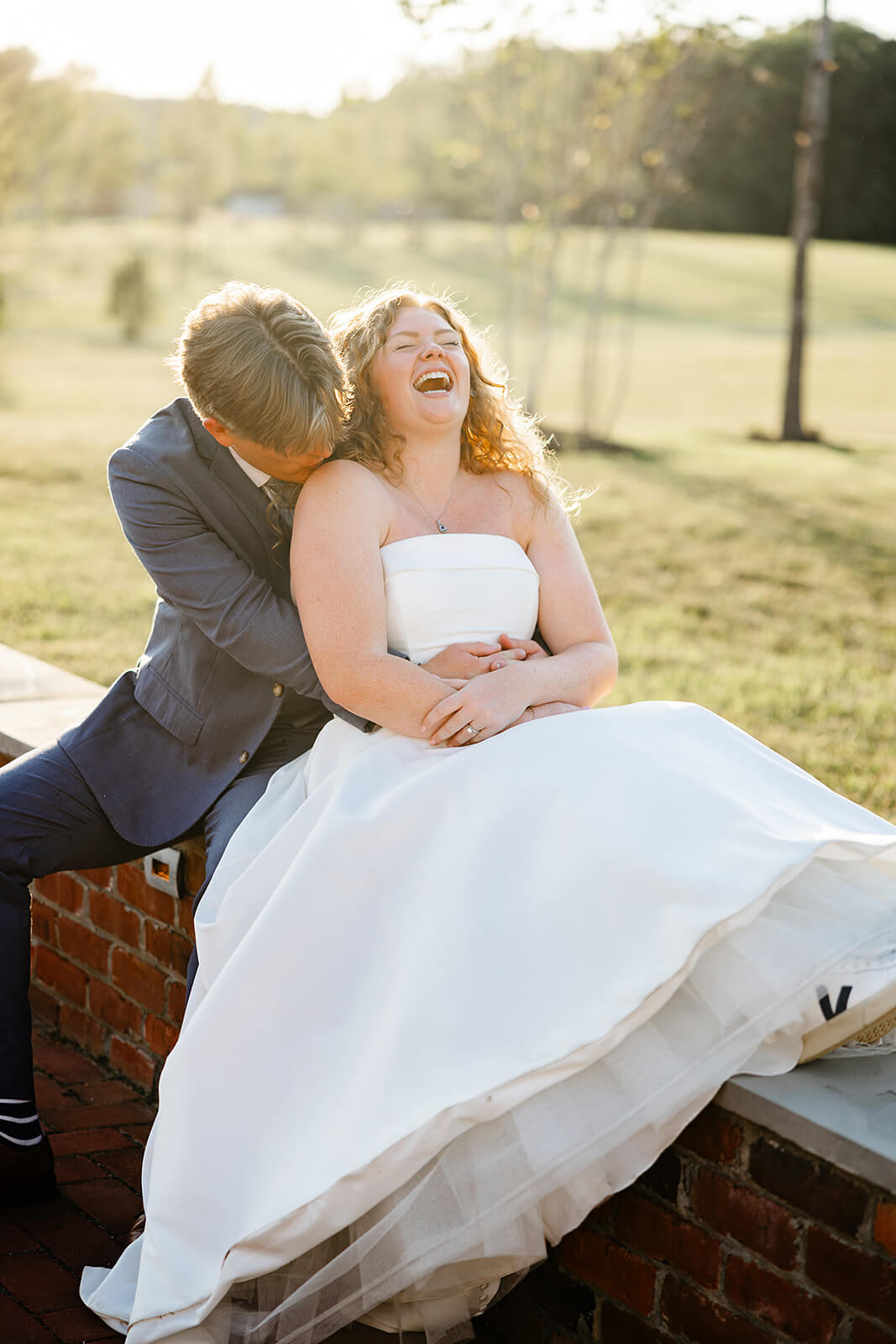 Rassawek Vineyard Spring Wedding Photography 31 groom hugging bride as he sits behind her on a short brick wall, the bride is laughing and the sun is setting behind them