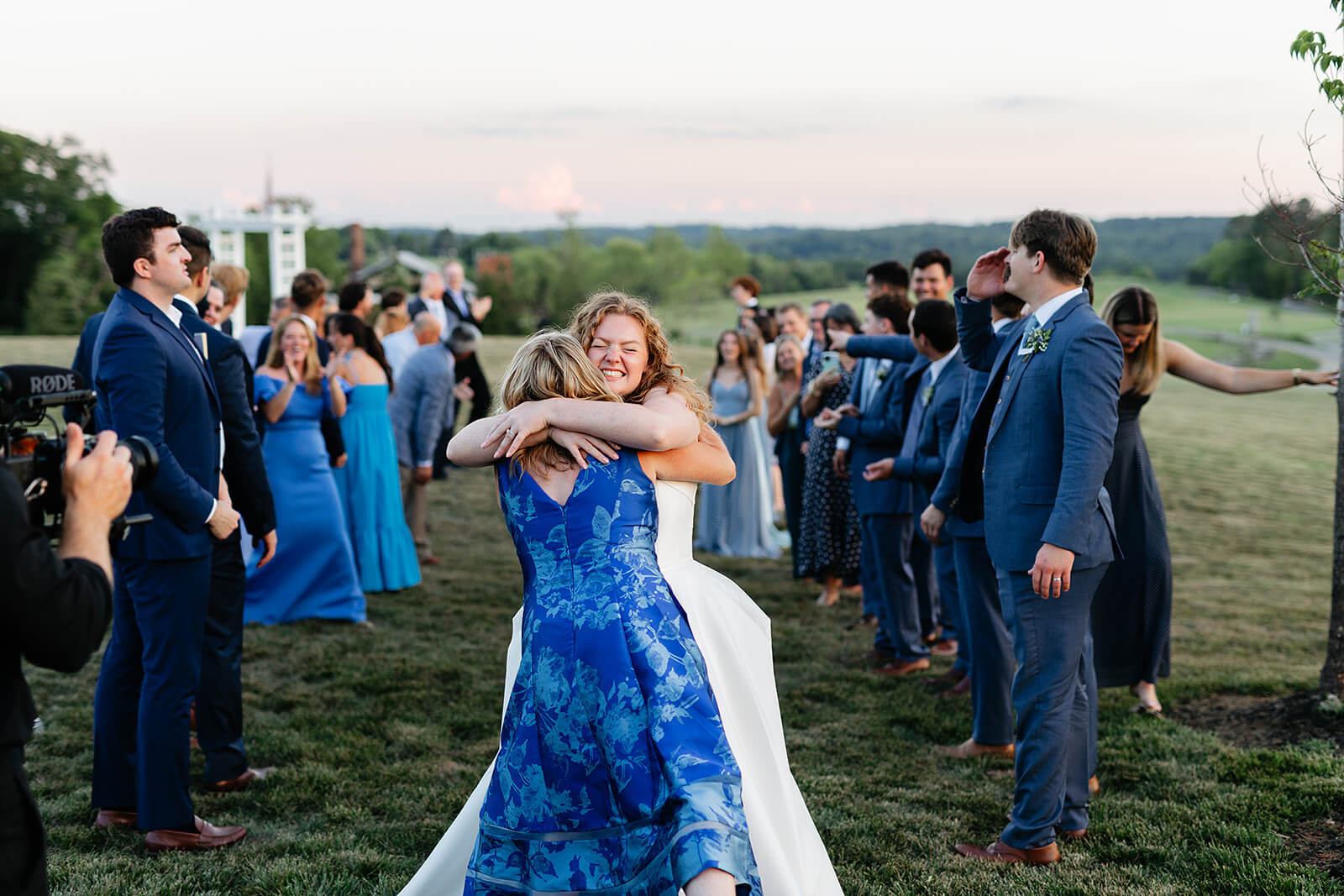 Rassawek Vineyard Spring Wedding Photography 47 bride hugging her mom outside during the exit, all the guests are standing on a large hilly lawn at sunset
