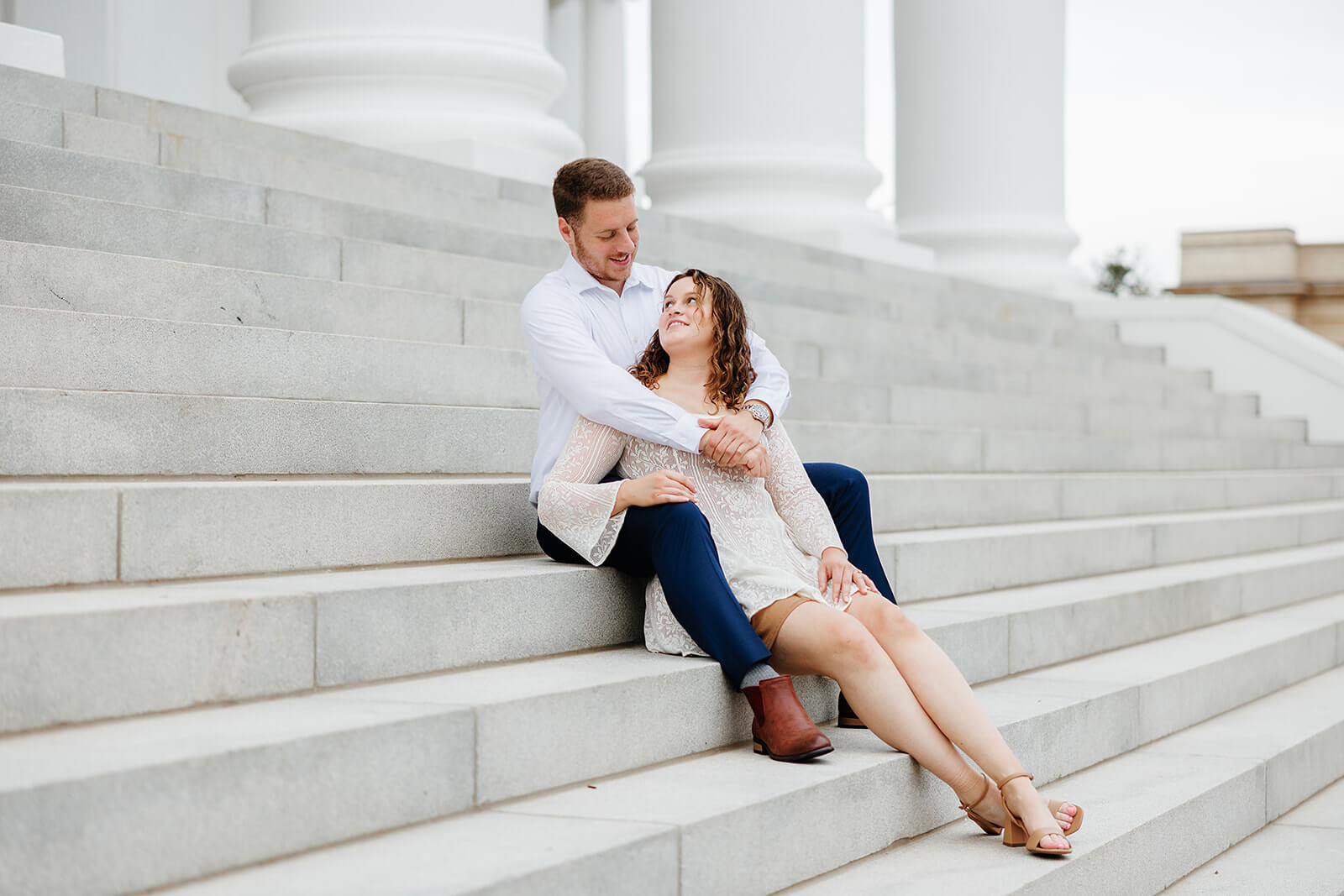 Virginia Capital Fall Engagement Photography 8 couple sitting on large white stairs, she is sitting between his legs and he is hugging her from behind, she is looking up at him
