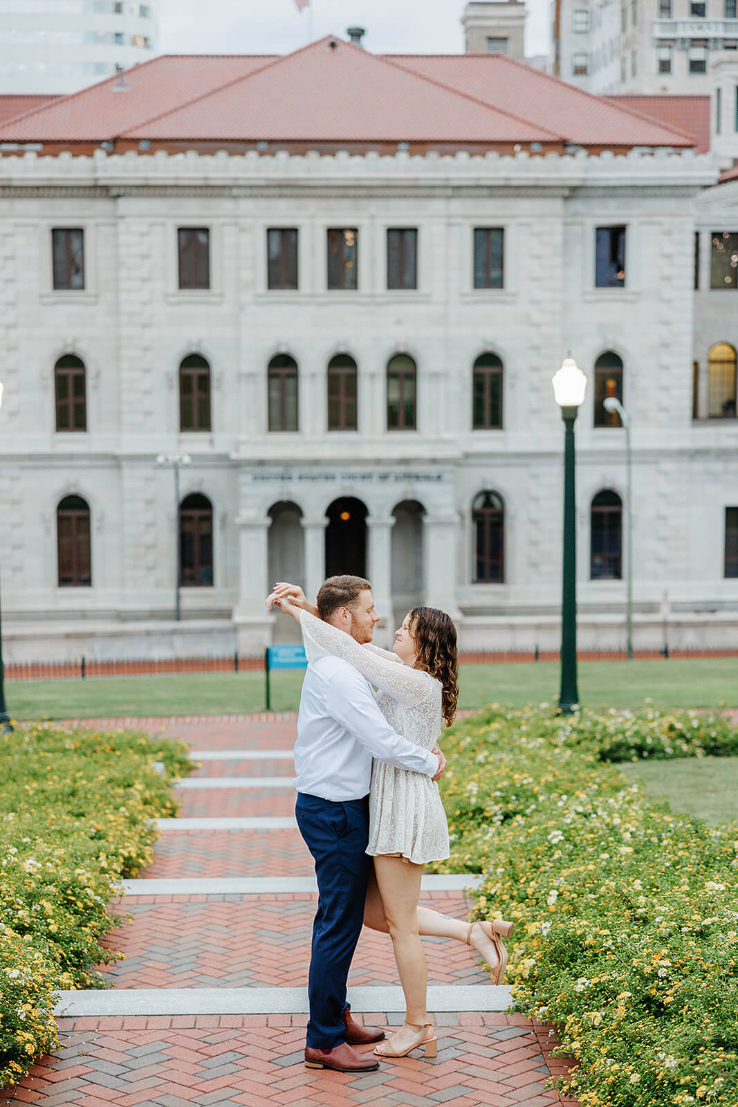 Virginia Capital Fall Engagement Photography 12 couple standing on brick path lined with yellow flowers and a white building with red roof in the background, they are hugging each other and he leg is popped in the air