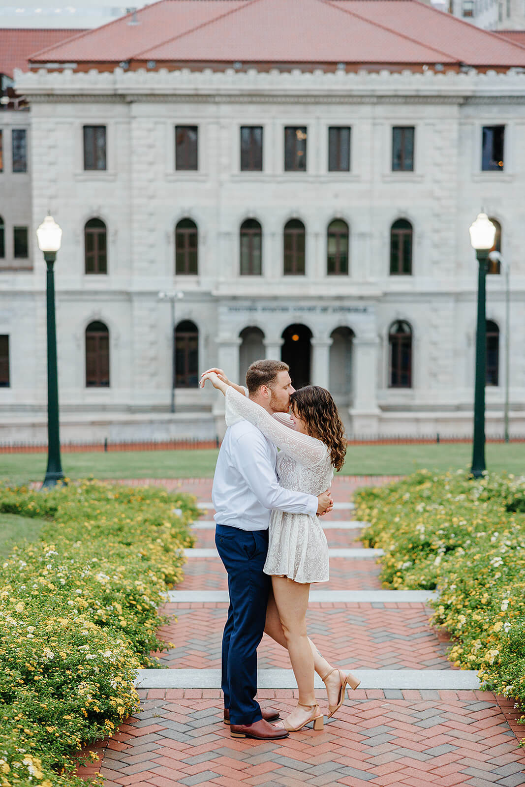 Virginia Capital Fall Engagement Photography 13 couple standing on brick path lined with yellow flowers and a white building with red roof in the background, they are hugging each other and he is kissing her on the forehead