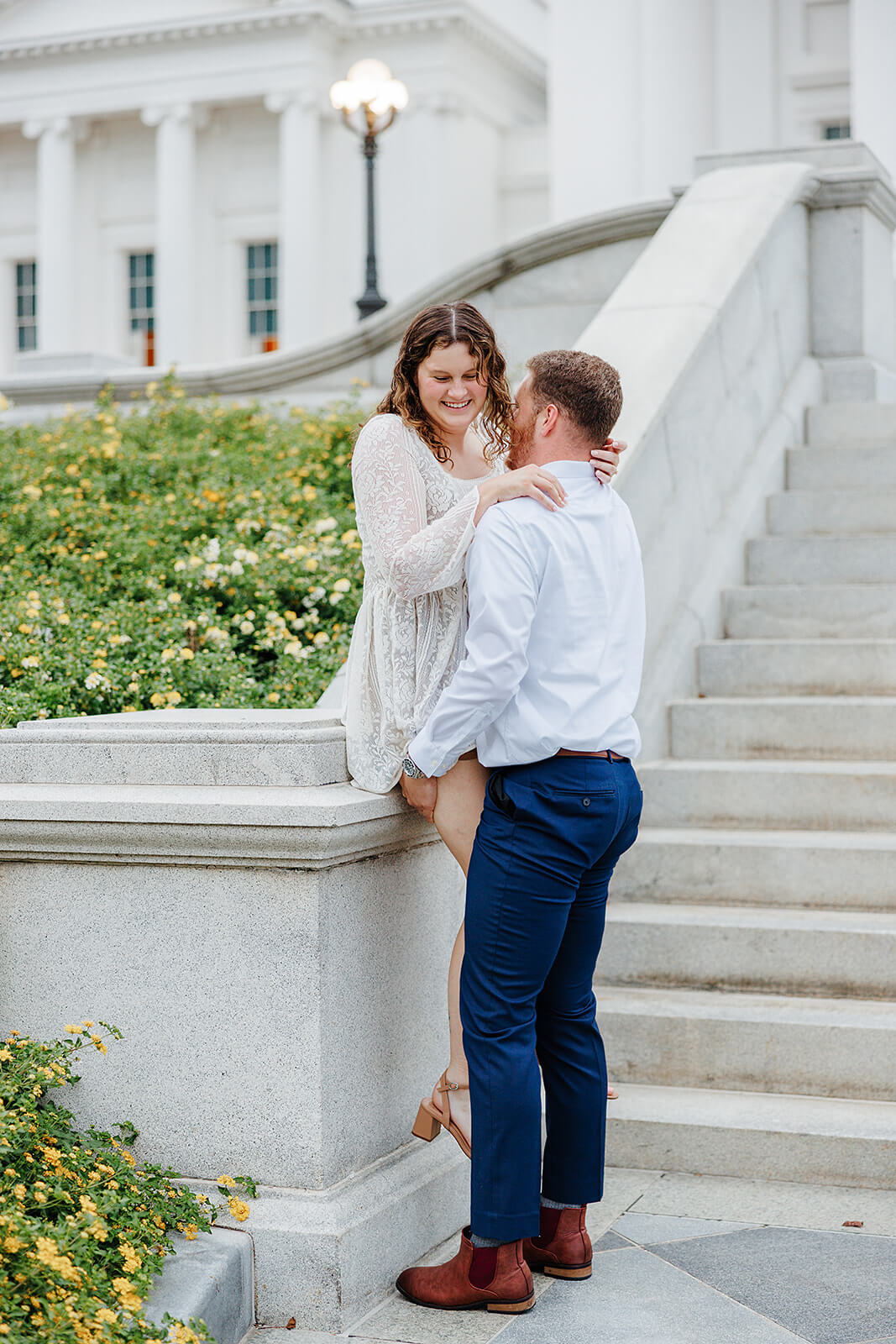 Virginia Capital Fall Engagement Photography 14 woman sitting on stone railing and her fiance is holding her up