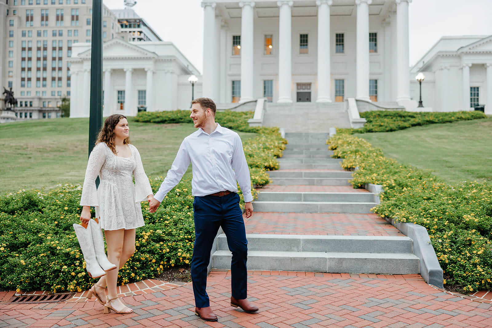 Virginia Capital Fall Engagement Photography 16 couple holding hands as they walk across brick path in front of large white building. she is holding a pair of tall white boots