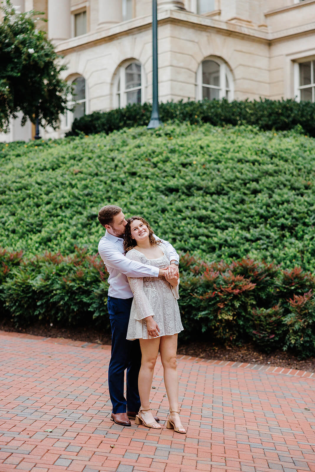 Virginia Capital Fall Engagement Photography 17 couple standing on brick path with a hill of bushes behind them, he is hugging her from behind