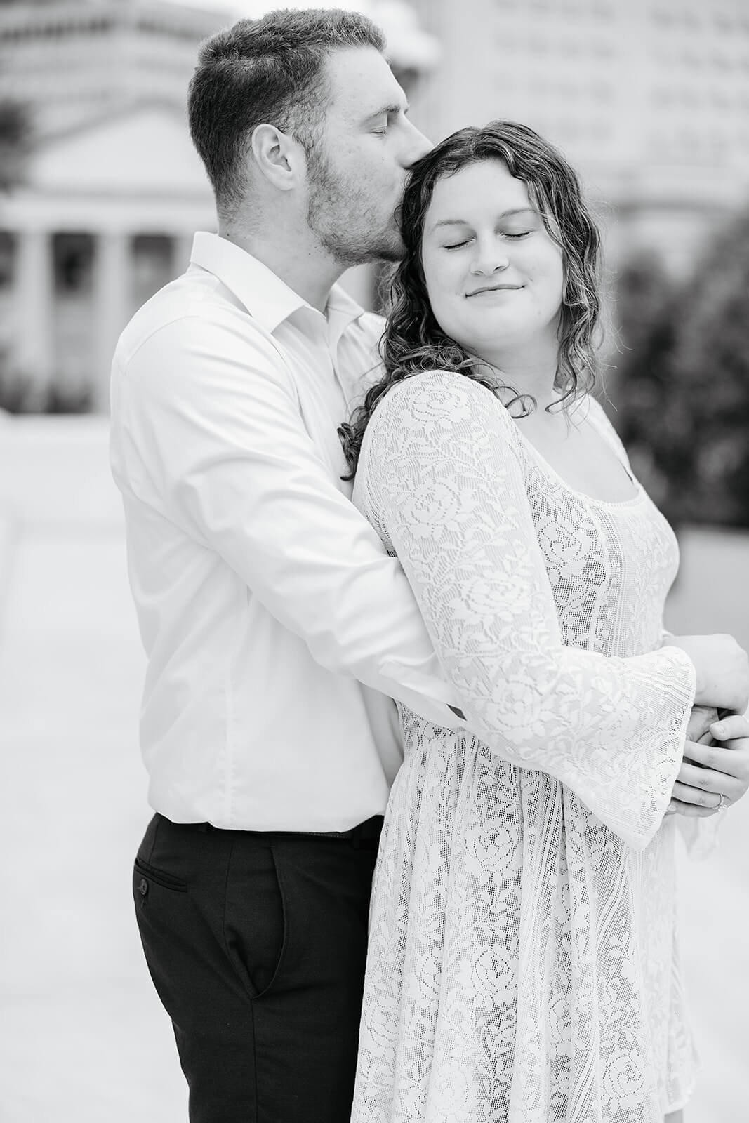 Virginia Capital Fall Engagement Photography 6 black and white photo of couple standing on large white stairs. he is hugging her from behind and kissing her on the head