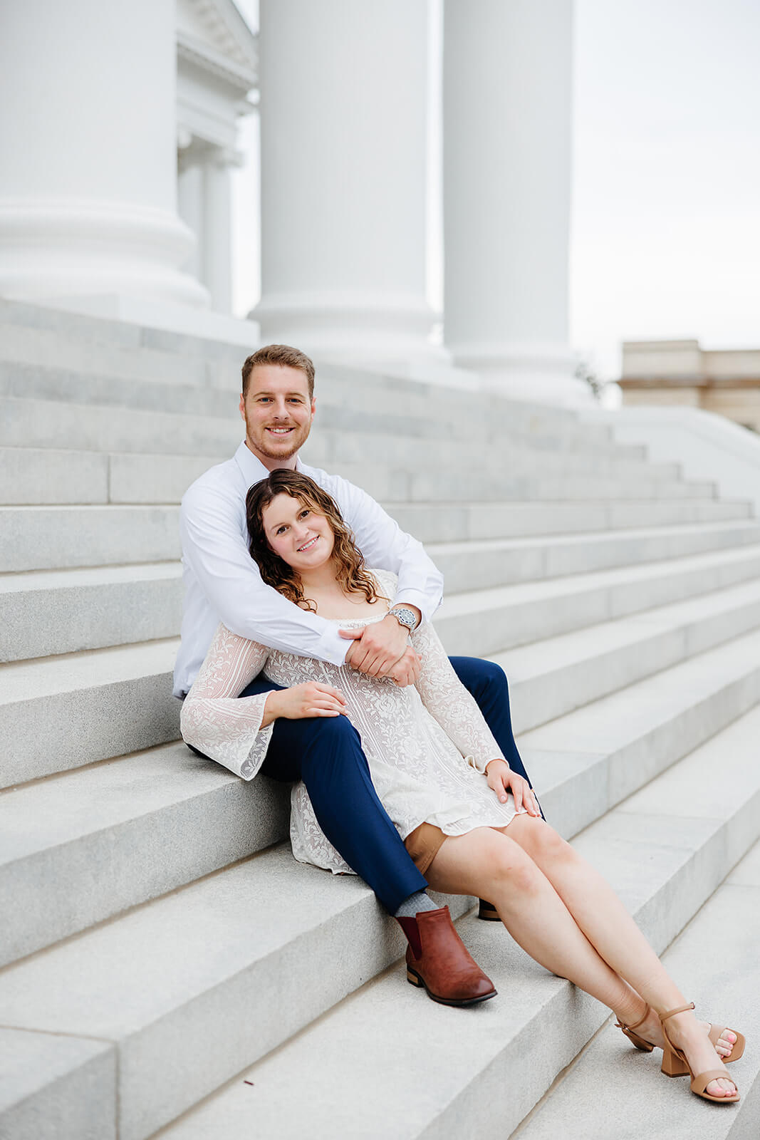 Virginia Capital Fall Engagement Photography 7 couple sitting on large white stairs, she is sitting between his legs and he is hugging her from behind